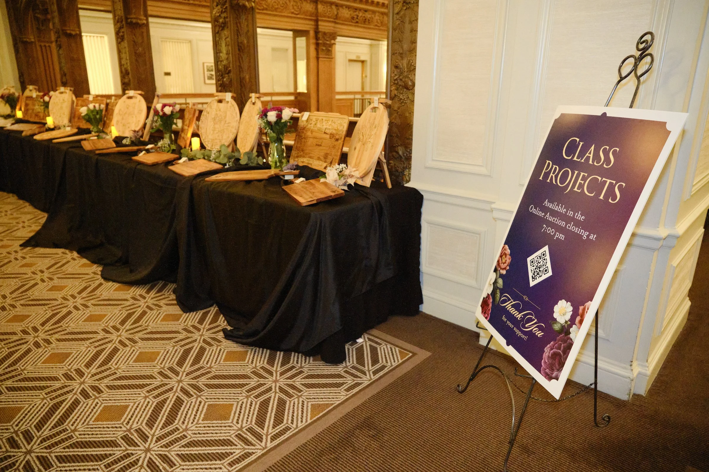 A table covered with a black cloth holding wooden plaques, vases with flowers, and small candles, set in a decorated indoor venue. To the right, a sign on an easel says 'Class Projects,' with details about an online auction ending at 7:00 pm, and a t