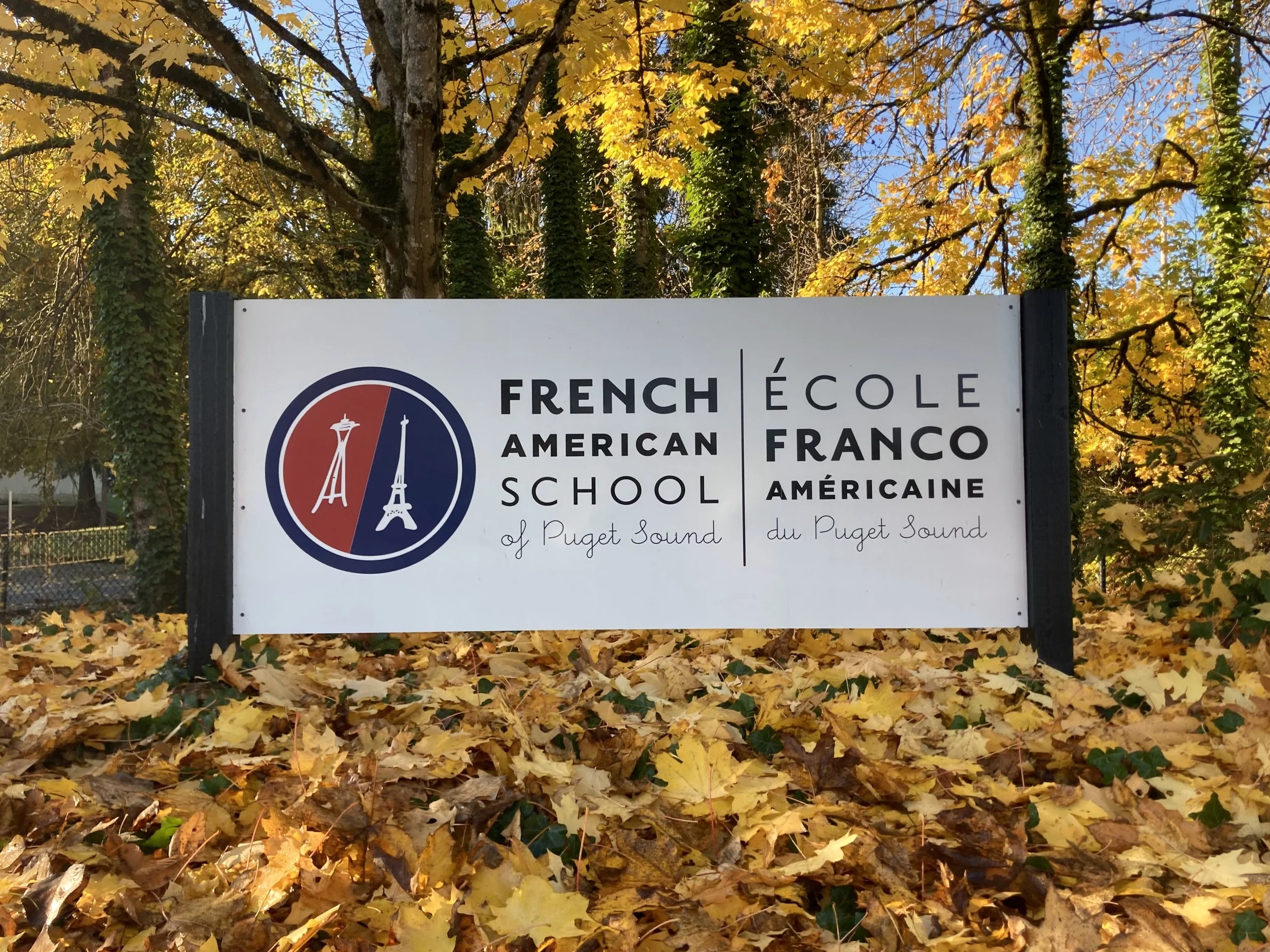 A sign for a bilingual school called 'French American School of Puget Sound' with a background of trees with yellow autumn leaves and fallen leaves on the ground.