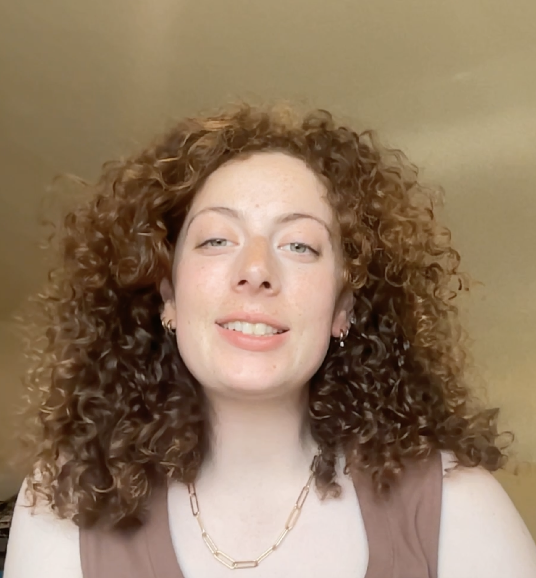 Close-up of a woman with curly red hair, smiling, wearing a gold chain necklace and earrings, and a white sleeveless top.