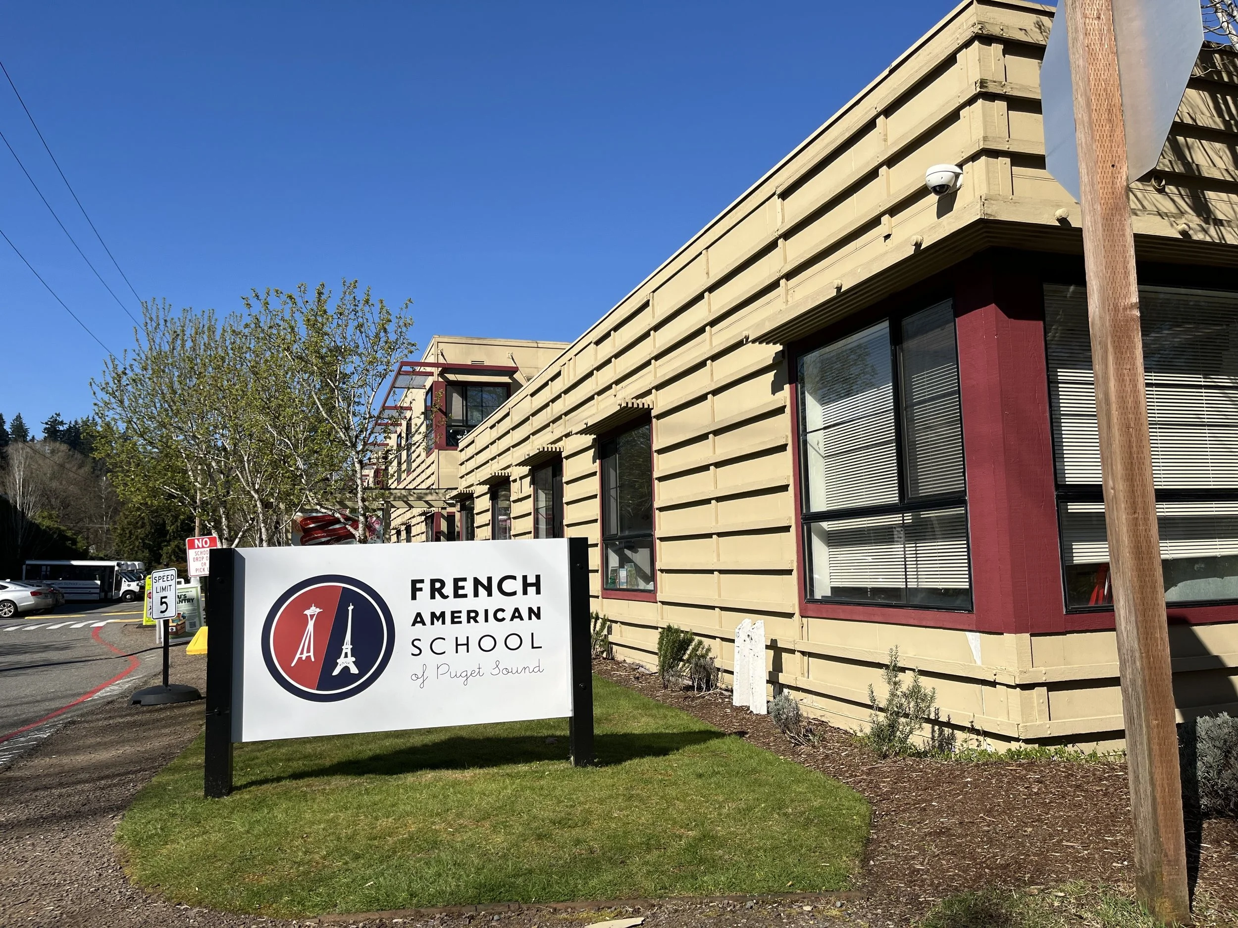 Exterior view of a building with beige siding and red accents, a sign for French American School of Puget Sound, and a security camera on the corner, with blue sky and trees in the background.