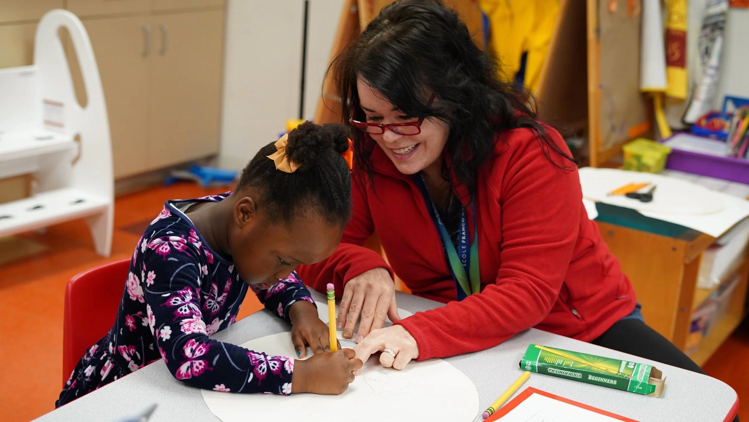 A woman with black hair and red glasses smiling while sitting at a table with a young girl who is drawing with a yellow crayon. The girl has her hair tied back with a beige bow and is wearing a navy blue dress with pink and white floral patterns. The background shows shelves with art supplies and a small white chair.