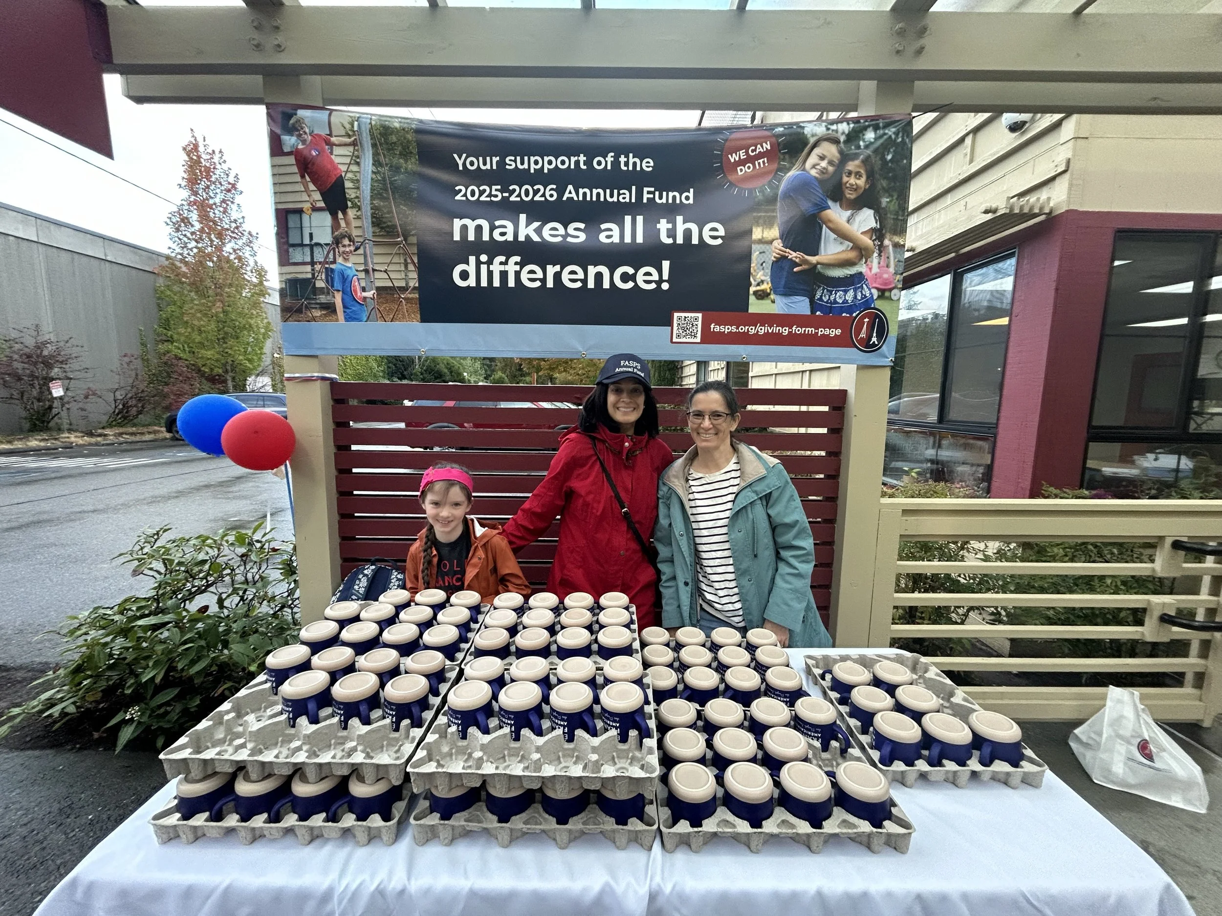People smiling standing behind a table with numerous coffee cups, outside near a building with a banner supporting an annual fund campaign.