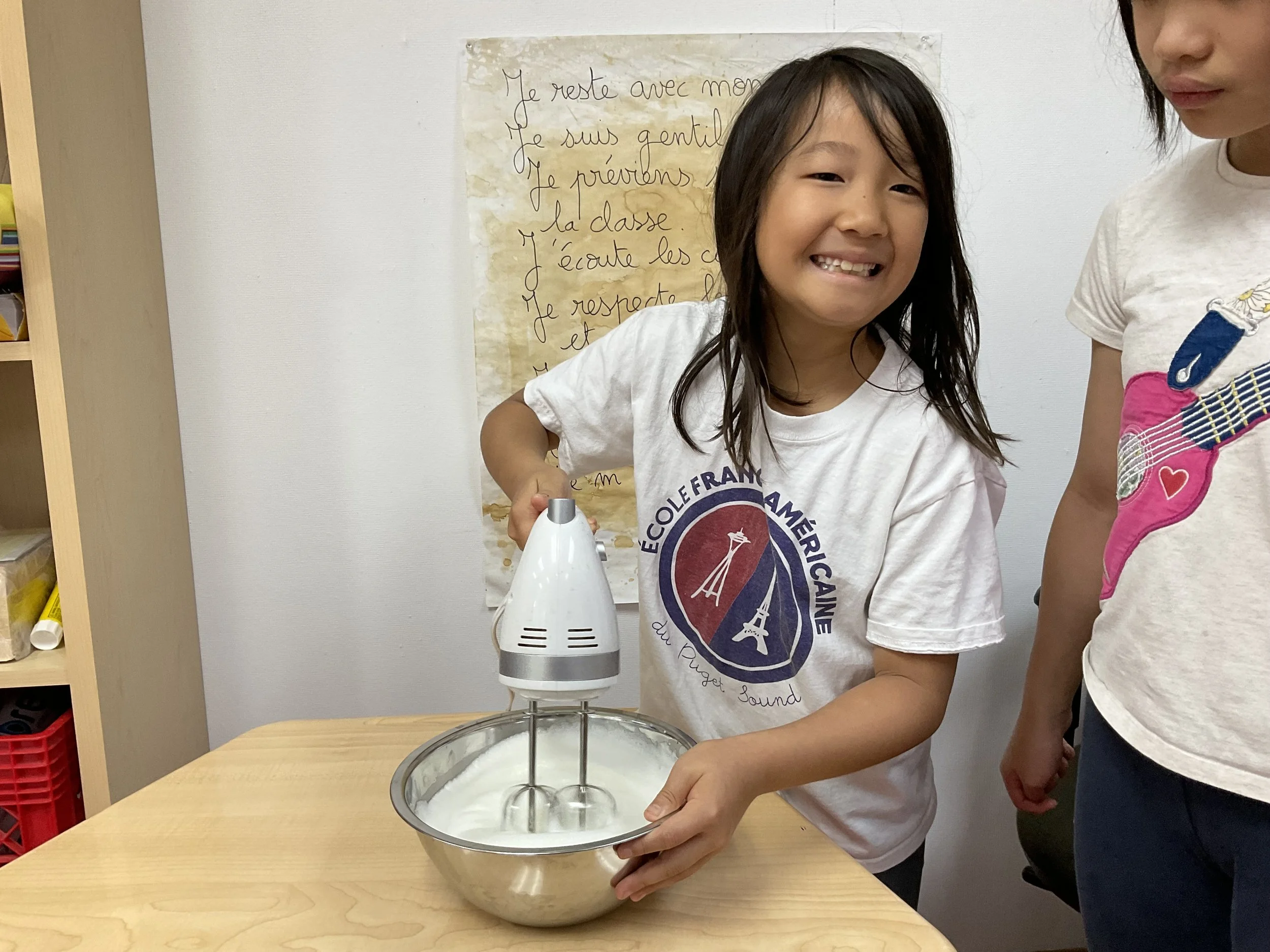 A young girl smiling while holding a mixing bowl on a table, with a shirt that has a logo and text in French. There is an anemometer in the bowl. A poster with French writing is on the wall behind her.