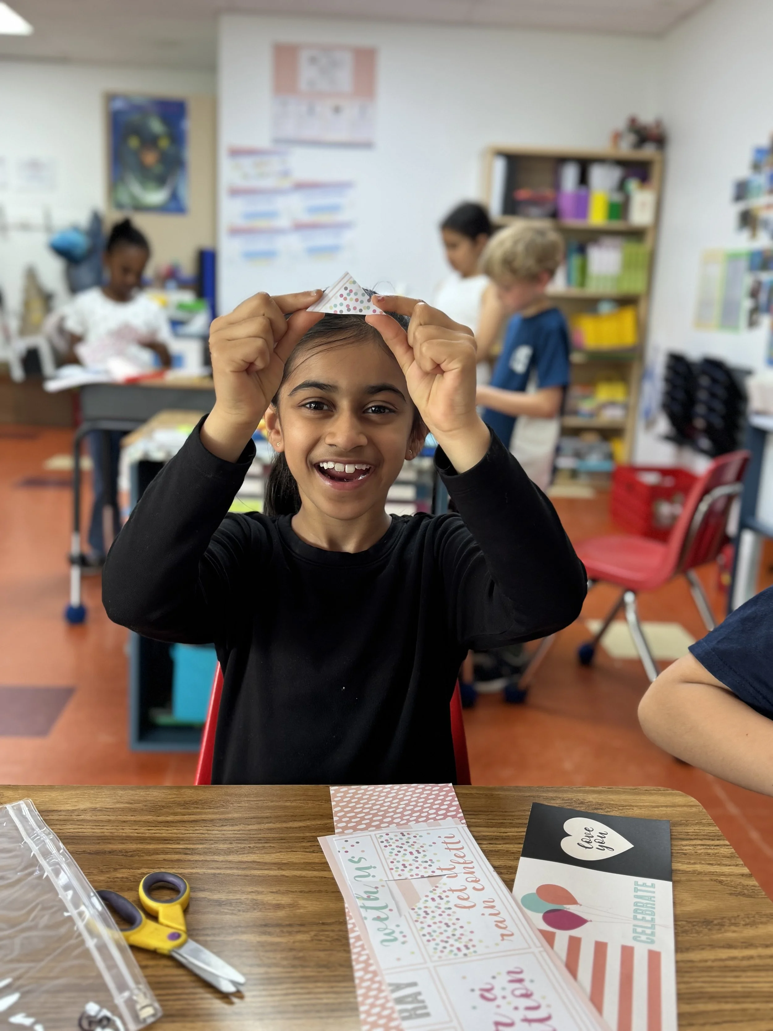 A young girl in a classroom is holding a small polka dot paper hat above her head, smiling at the camera. In the background, other children are engaged in activities, with classroom supplies and bookshelf visible. On the table in the foreground, there are scissors, greeting cards, and craft materials.