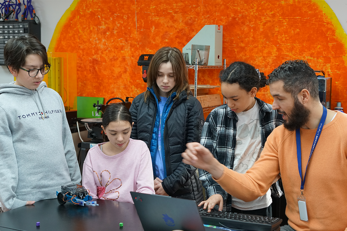 An instructor demonstrating robotics to a group of children in a classroom or makerspace, with a small robot on the table.