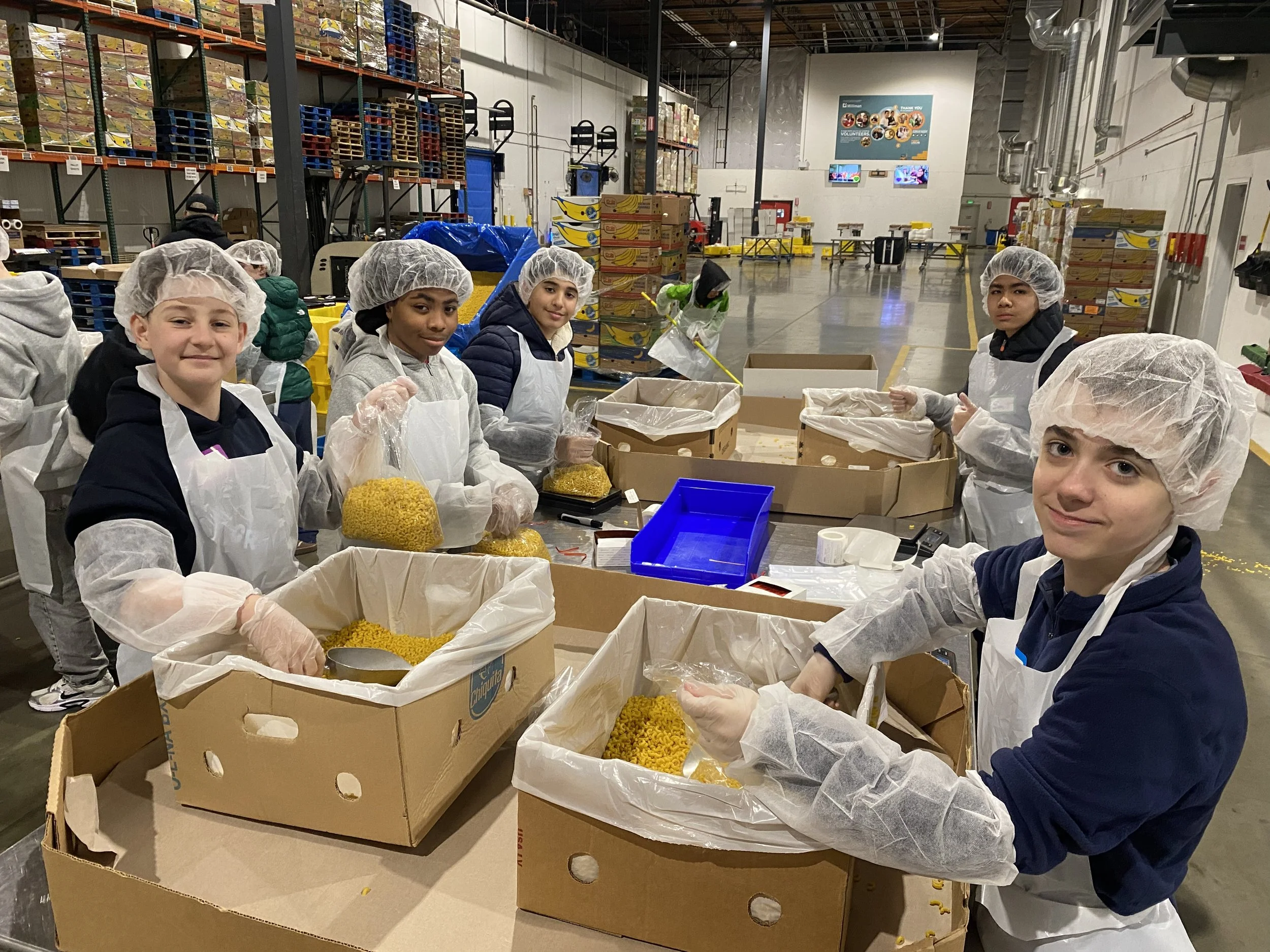 Children wearing hairnets and gloves working in a food packaging facility, packing noodles into boxes, with shelves of boxes and pallets in the background.