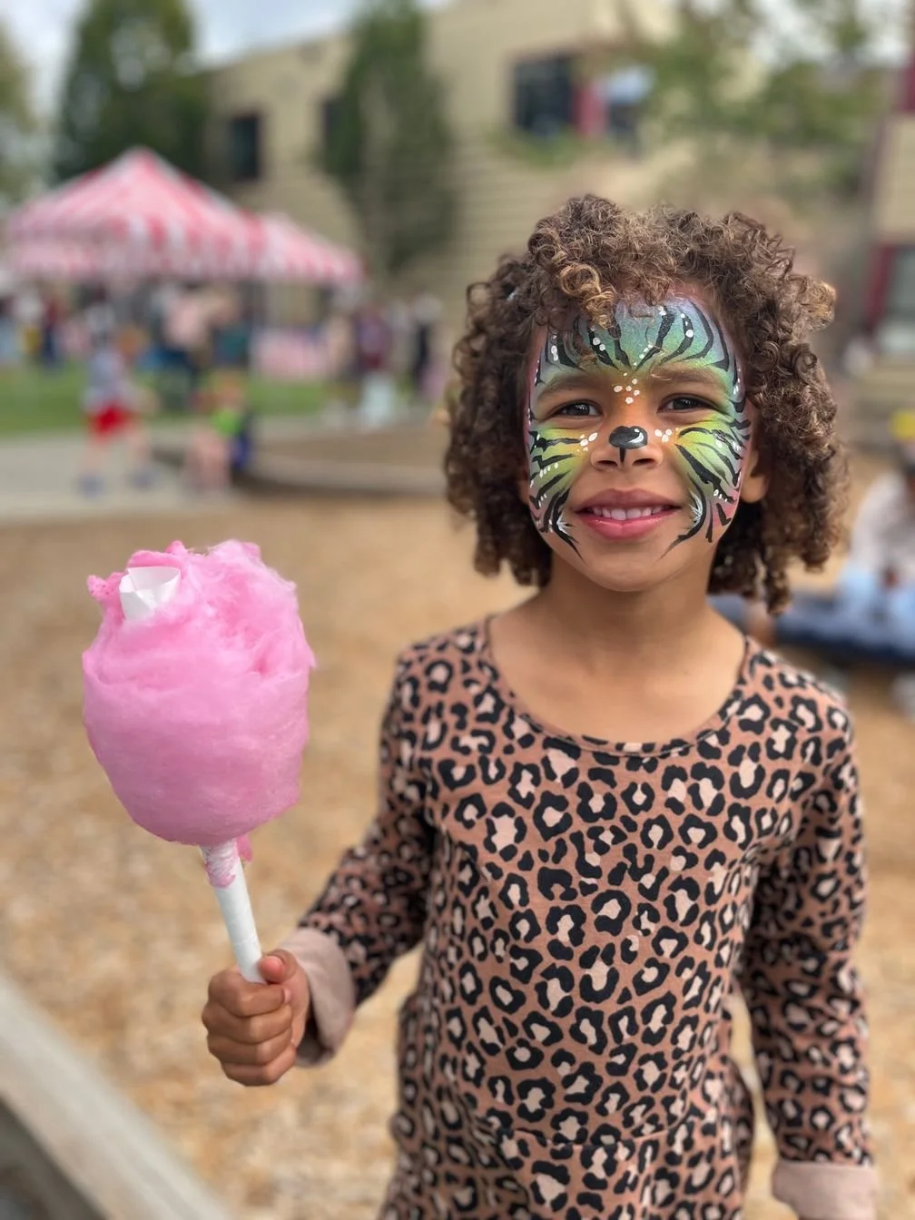 A young girl with face paint resembling a colorful animal, wearing a leopard print dress, holding pink cotton candy at an outdoor event with a blurred background of people, buildings, and a red and white striped tent.