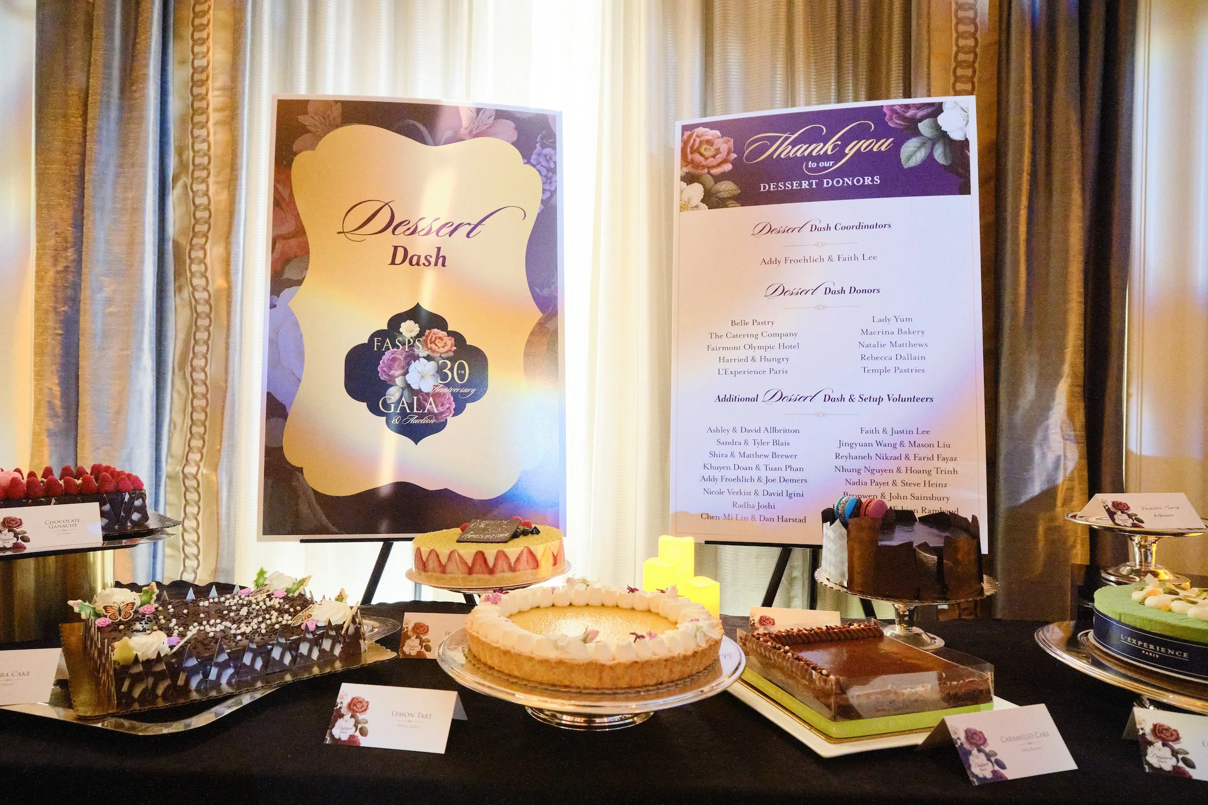 A dessert table with various cakes and sweets, two large decorative signs for an event celebrating a 30th anniversary gala, floral decorations, and a backdrop of curtains.