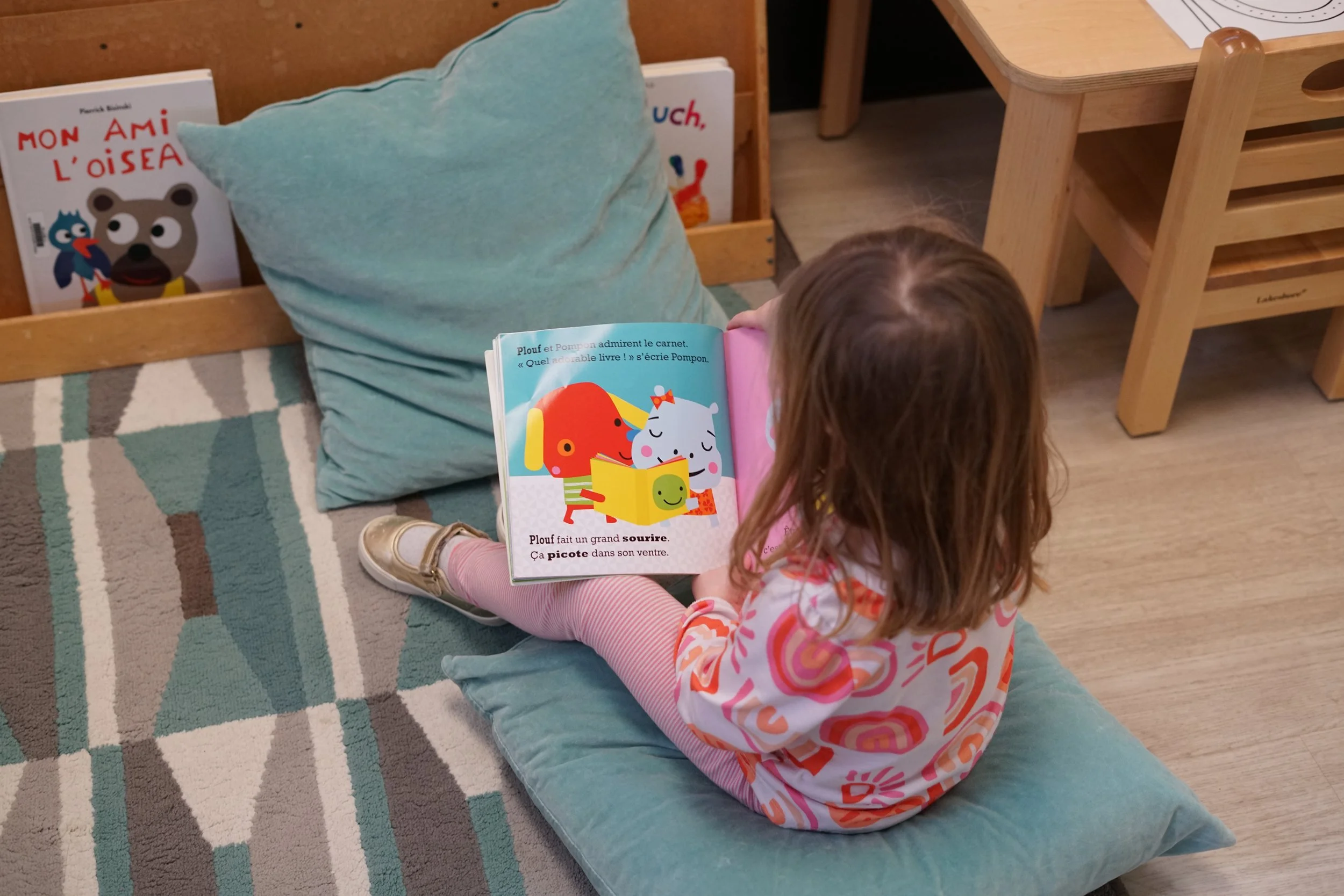 A young girl with curly hair, wearing a colorful patterned shirt and pink striped pants, sitting on a blue cushions reading a children's book with colorful illustrations of animals.