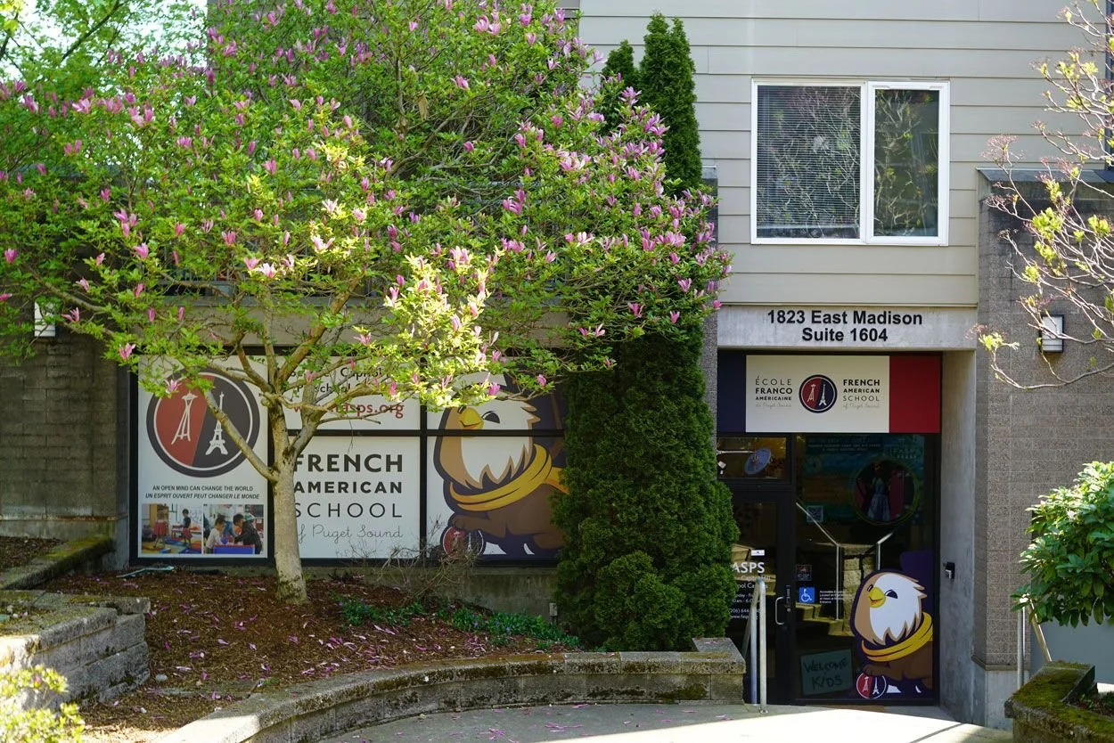 Entrance to French American School with a colorful sign, a tree with pink flowers, and the building address 1823 East Madison Suite 1604.