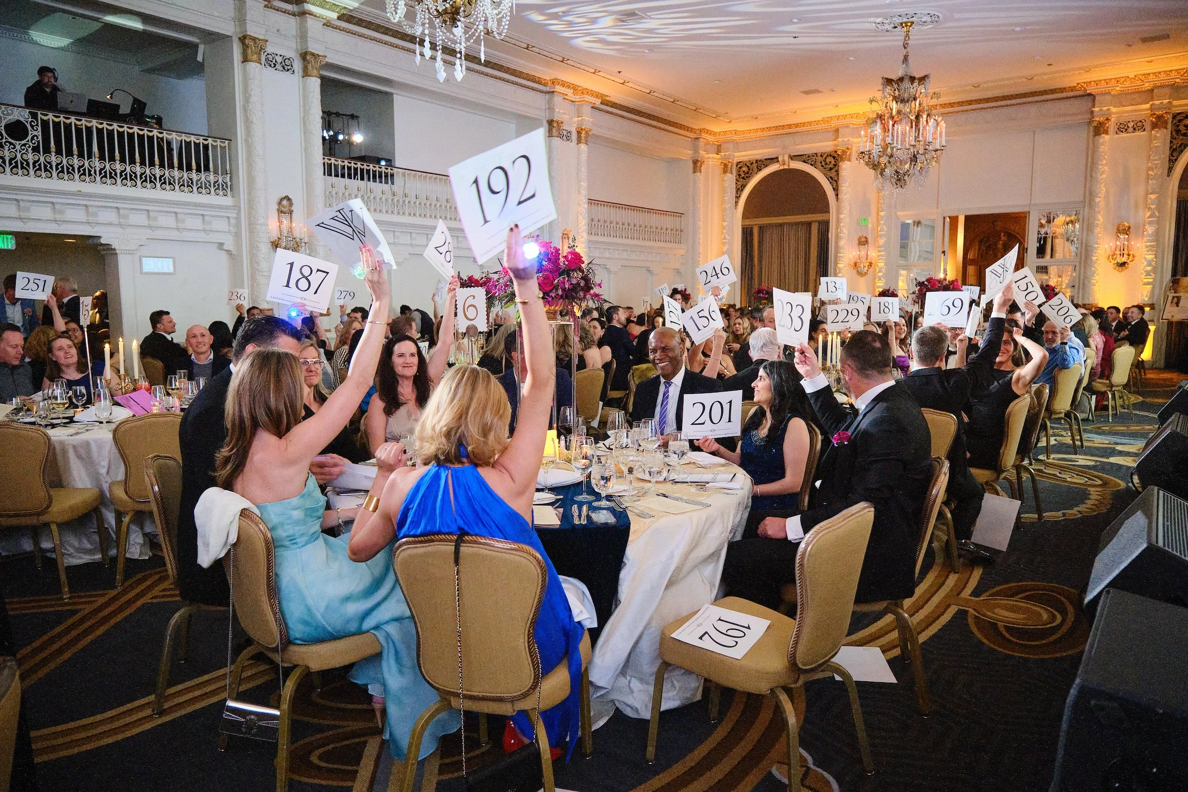 A formal event or auction in a luxurious ballroom with chandeliers, ornate decor, and many people seated at tables. Attendees hold up numbered paddles, possibly bidding.