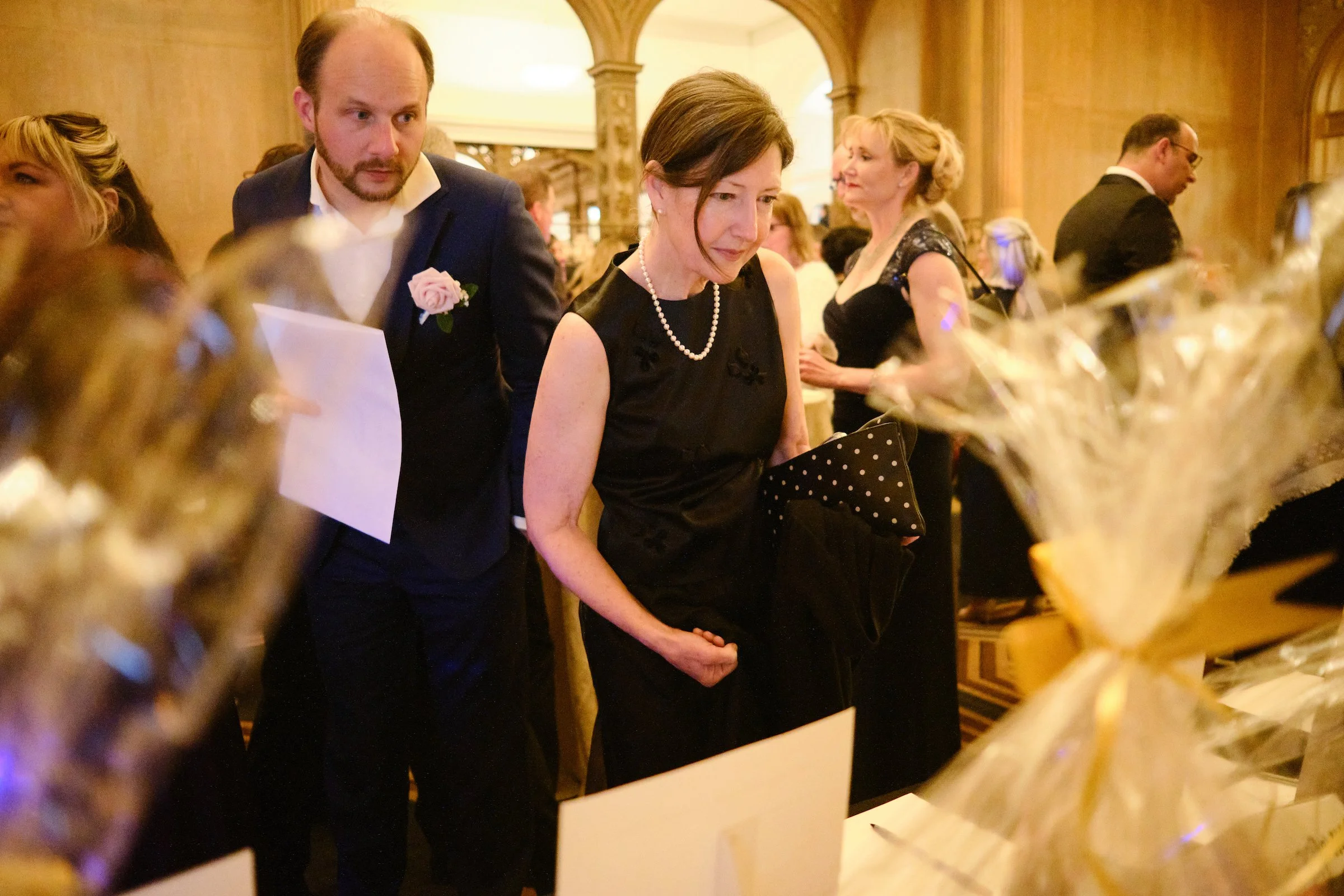 People in formal attire at an indoor event, possibly a wedding reception, with golden decorations and floral arrangements in the foreground.