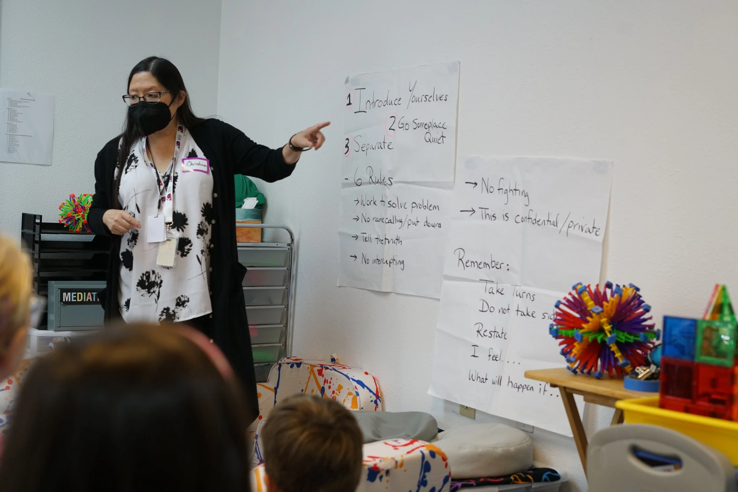 A woman wearing a black mask and glasses, pointing at a whiteboard with instructions, standing in a classroom or workshop setting with children sitting in front of her. The whiteboard contains a list of rules and guidelines for a group activity.