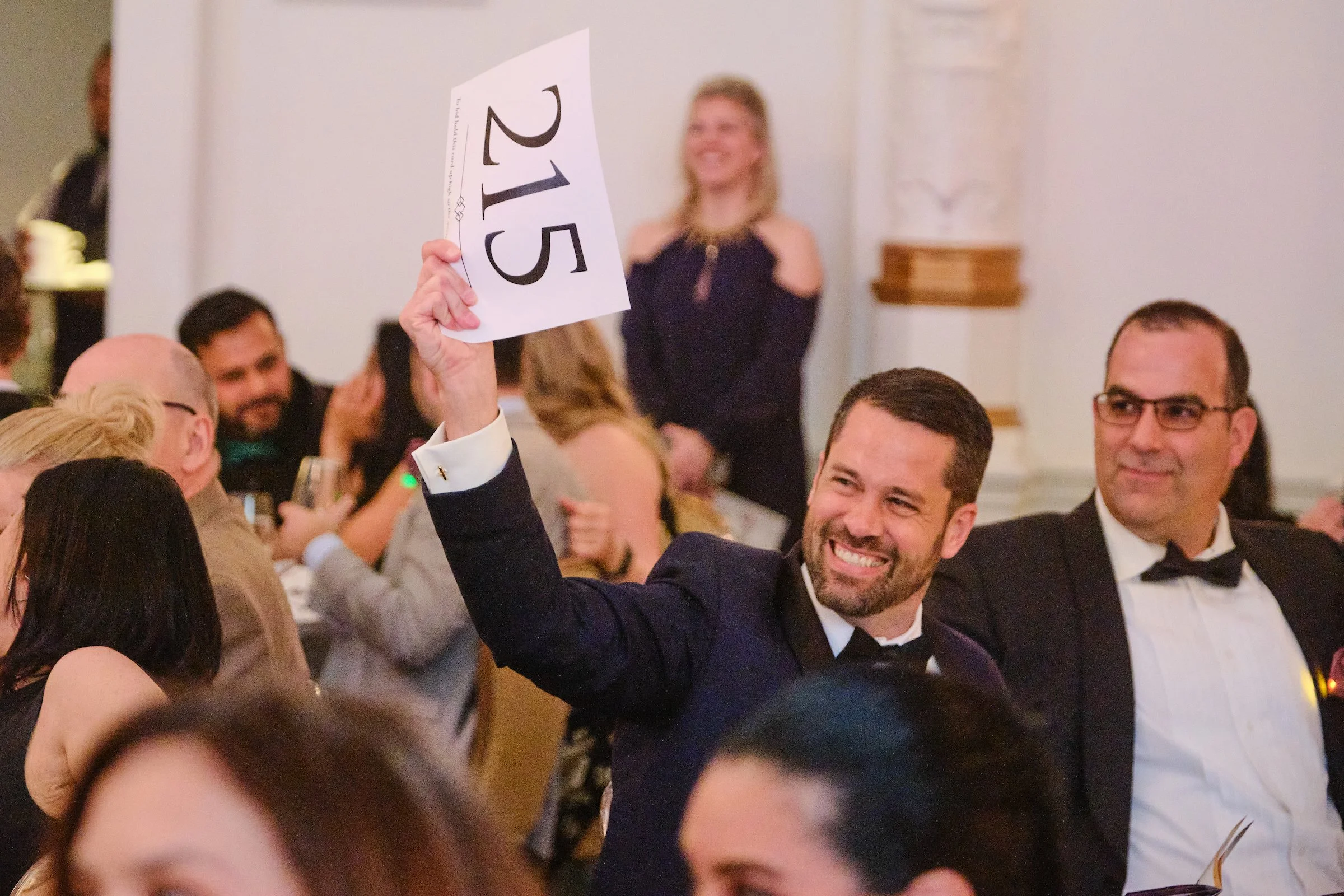 A man dressed in a tuxedo and bow tie is smiling and raising a card with the number 215 at a formal event or auction. There's a crowd of people around him, some wearing suits and dresses, and a woman in a black dress and necklace is visible in the ba