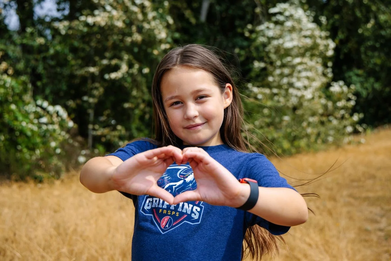 A young girl with long brown hair smiling at the camera, making a heart shape with her hands in an outdoor setting with trees and grass.