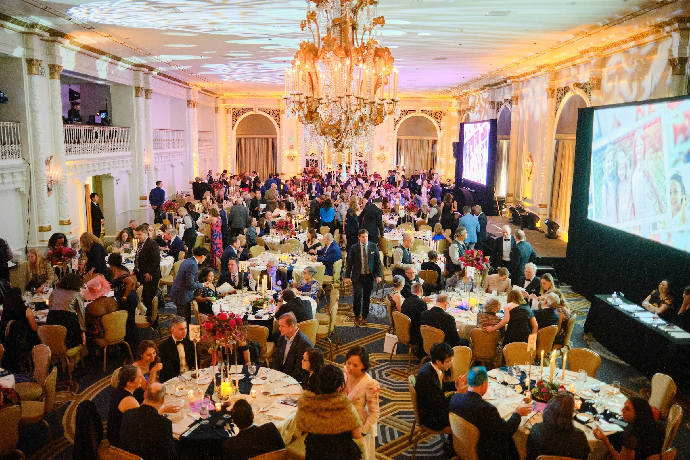 A large, elegant banquet hall filled with many formally dressed people seated at round tables decorated with pink floral centerpieces and lit candles. There is a large ornate chandelier hanging from the ceiling, and two big screens on the right displ