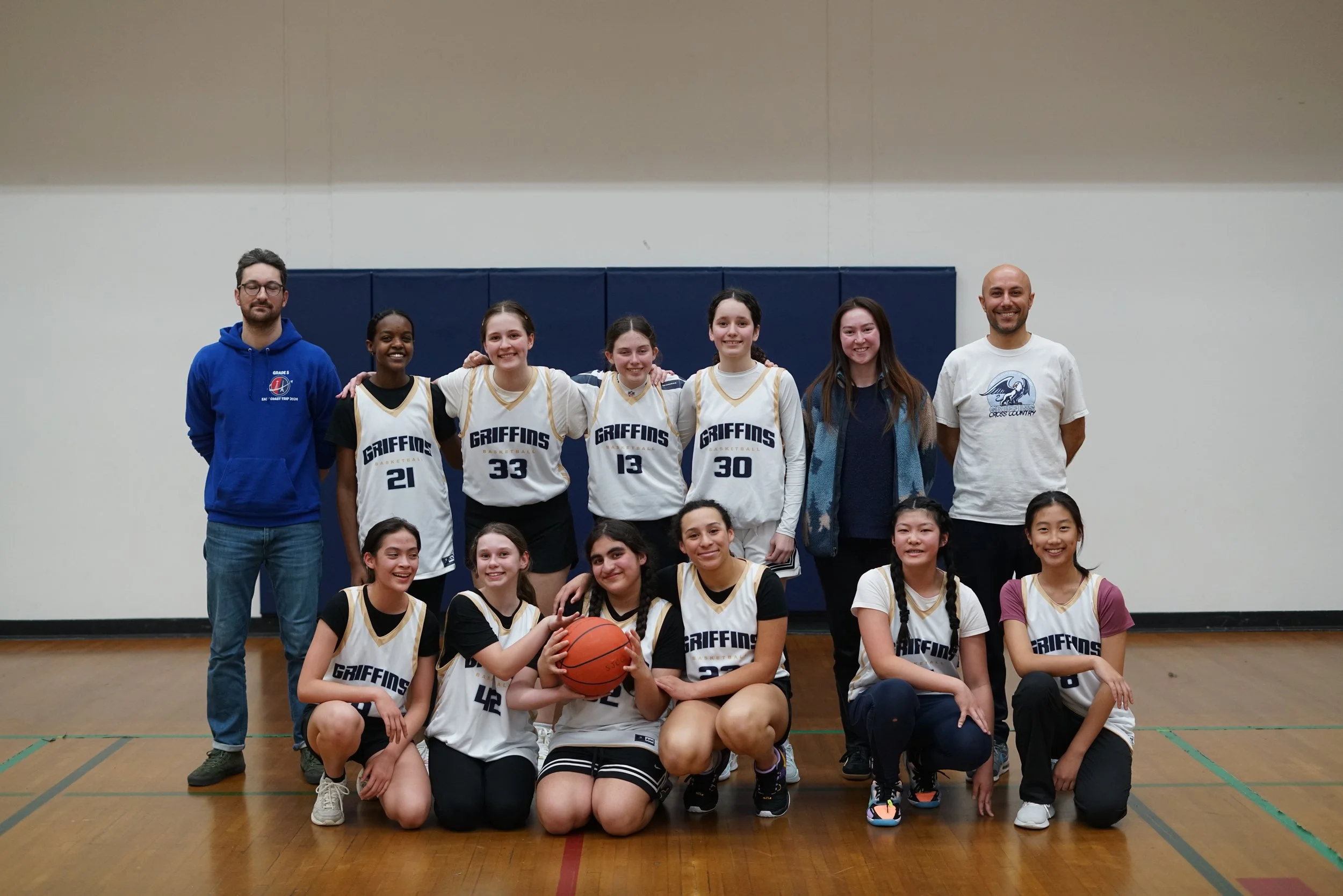 A girls basketball team and two coaches standing and sitting together in a gymnasium, smiling for the camera, with a blue padded wall behind them. The girls are wearing white jerseys with the team name 'Gryffins' and their jersey numbers, and one girl is holding a basketball. The coaches are wearing casual clothing, one in a blue hoodie and the other in a white T-shirt.