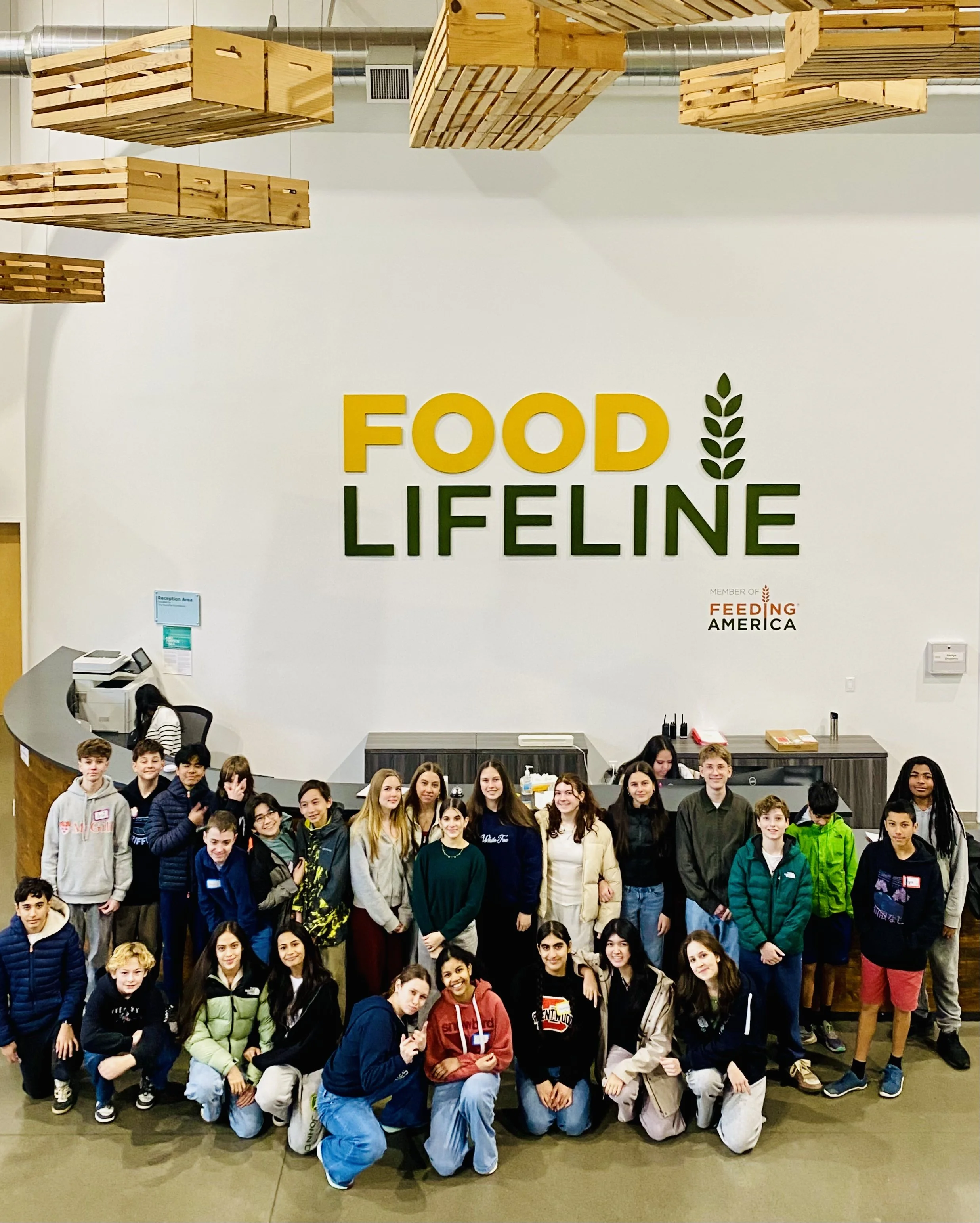 Group of children and teenagers posing for a photo in front of a large sign that says 'Food Lifeline' with a green leaf graphic. The group is inside a building, possibly a food bank or community center, with a reception desk and wooden ceiling fixtures.