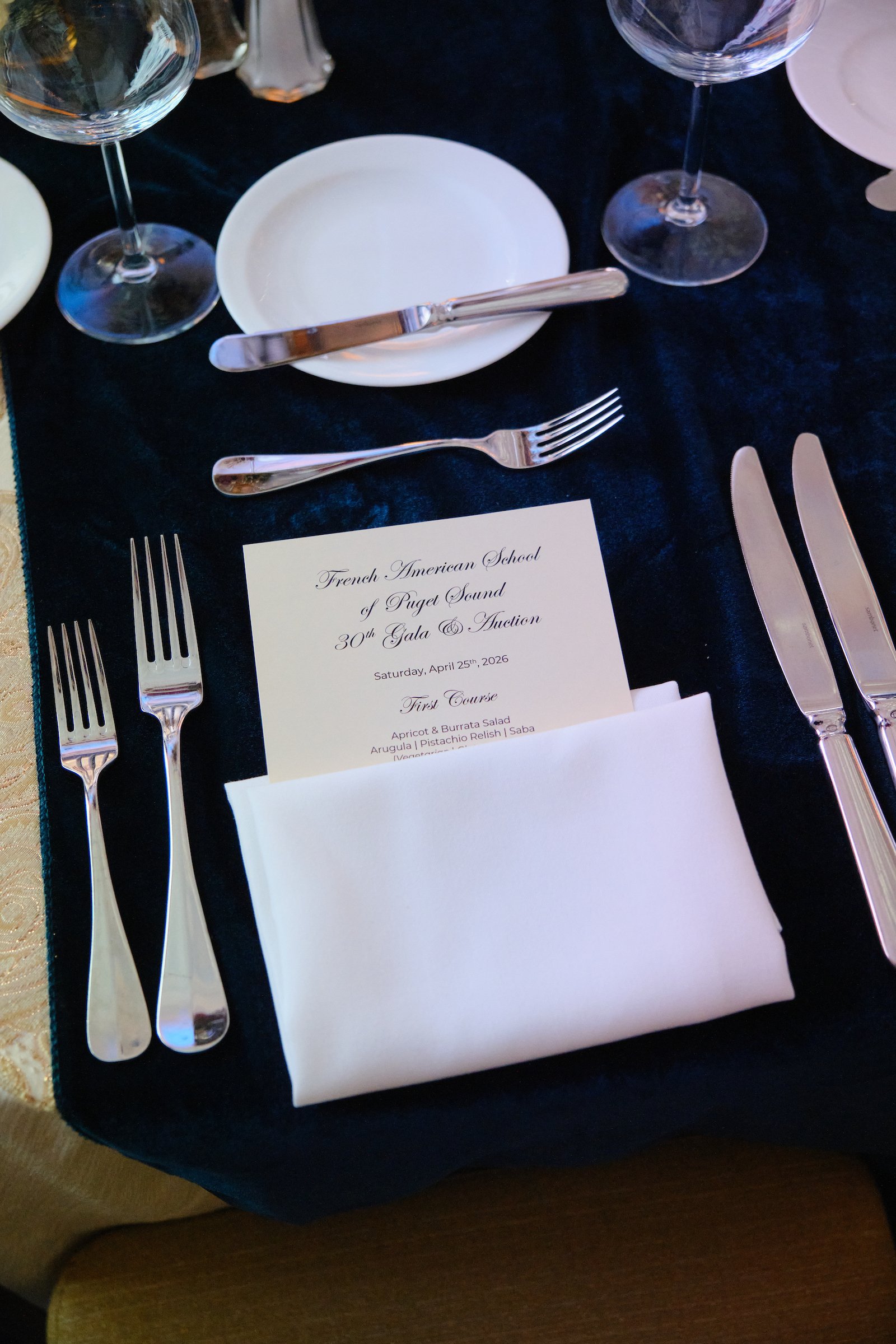 Table setting with two forks, two wine glasses, a knife, a small white plate, and a menu for the French American School of Puget Sound's 30th Gala and Auction, held on Saturday, April 25th, 2026.