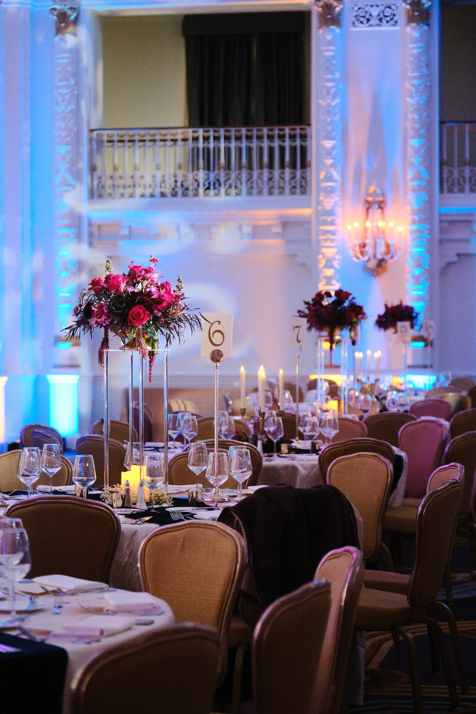 Elegant banquet hall decorated for a wedding reception with floral centerpieces, candles, and table settings, featuring a two-level venue with ornate architectural details and soft lighting.