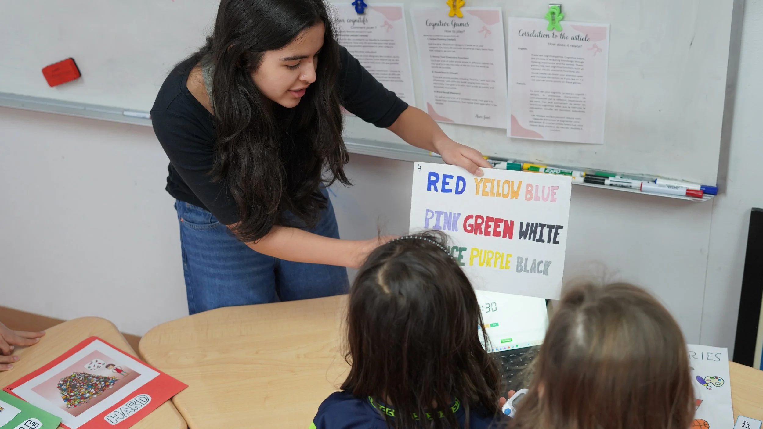 A woman is holding a colorful educational poster with color names written on it, in a classroom setting, with children seated in front of her.