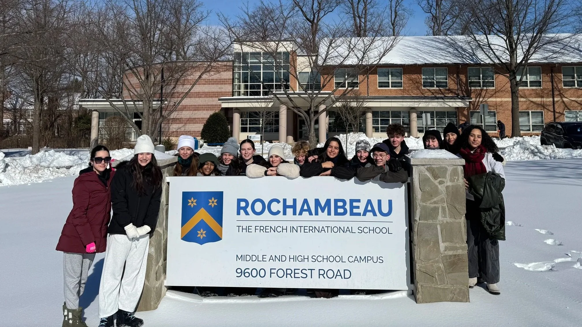 A group of diverse students standing outside in the snow, posing behind a sign for Rochambeau, the French International School. The school building is visible in the background with leafless trees and a clear blue sky.