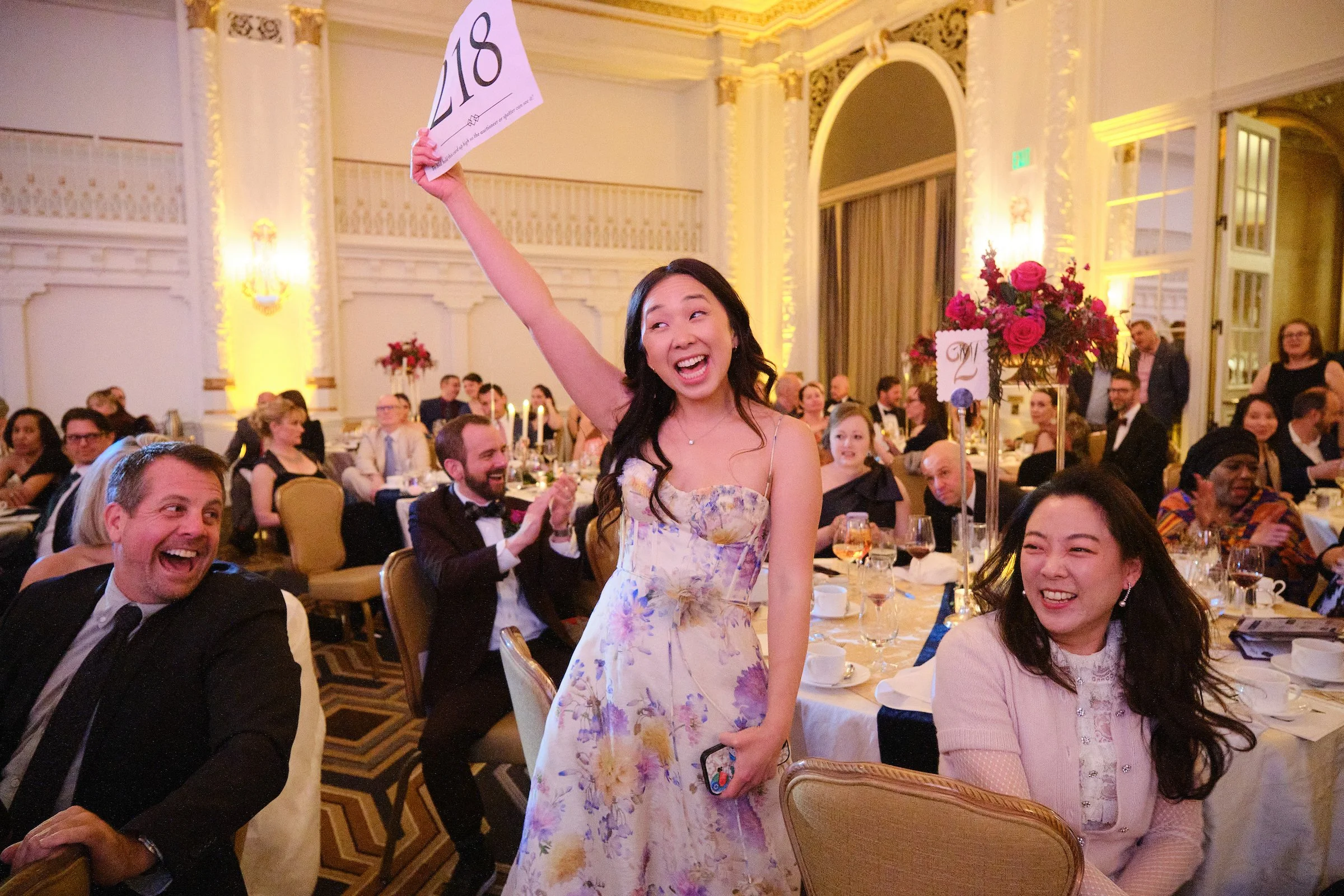 A woman in a floral dress is standing and smiling at a formal event, holding a sign that says '2018,' while guests seated at tables and clapping are visible in the background.