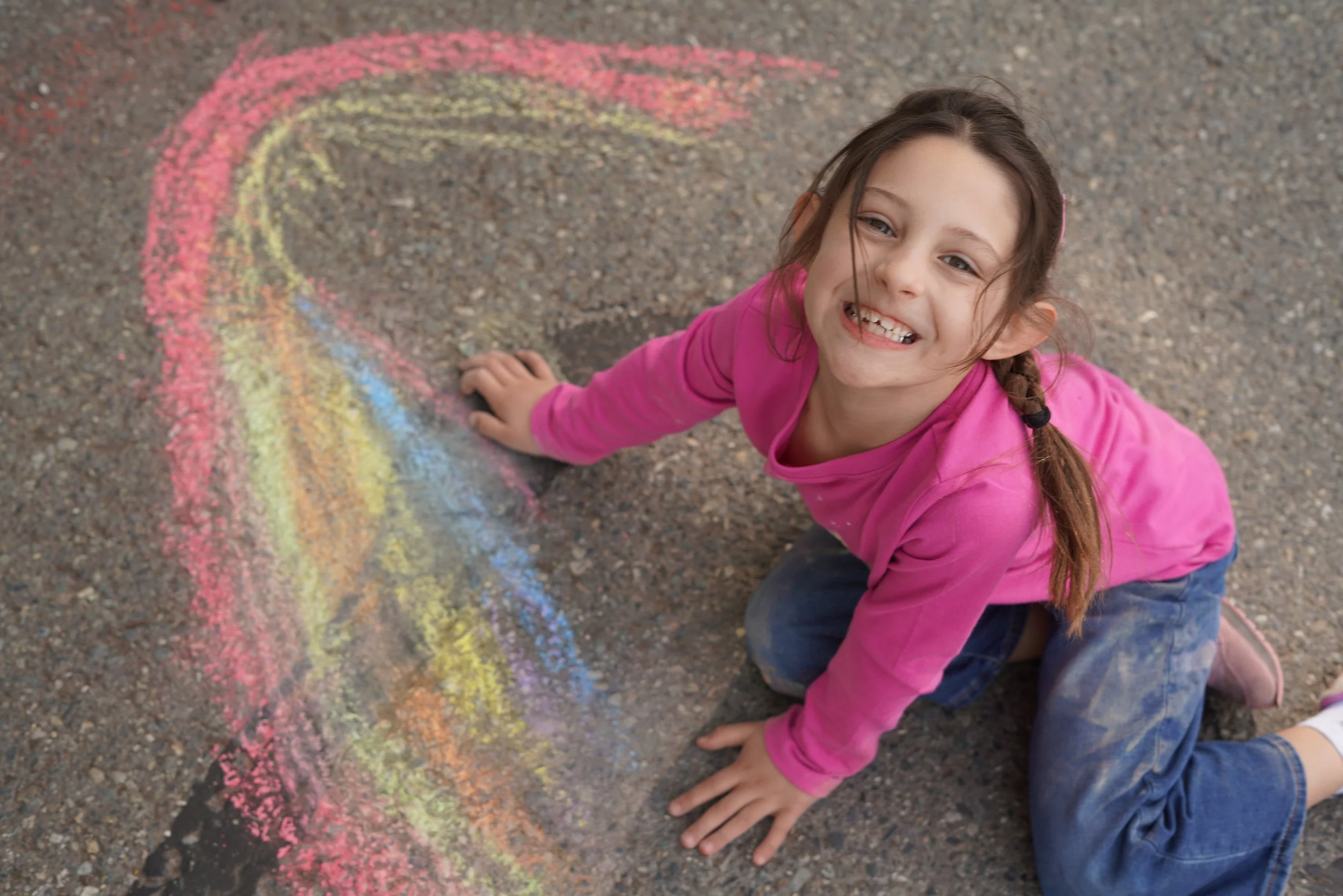 A young girl with brown hair in a braid, wearing a pink long-sleeve shirt and blue jeans, smiling and sitting on the ground next to a colorful chalk rainbow on asphalt.