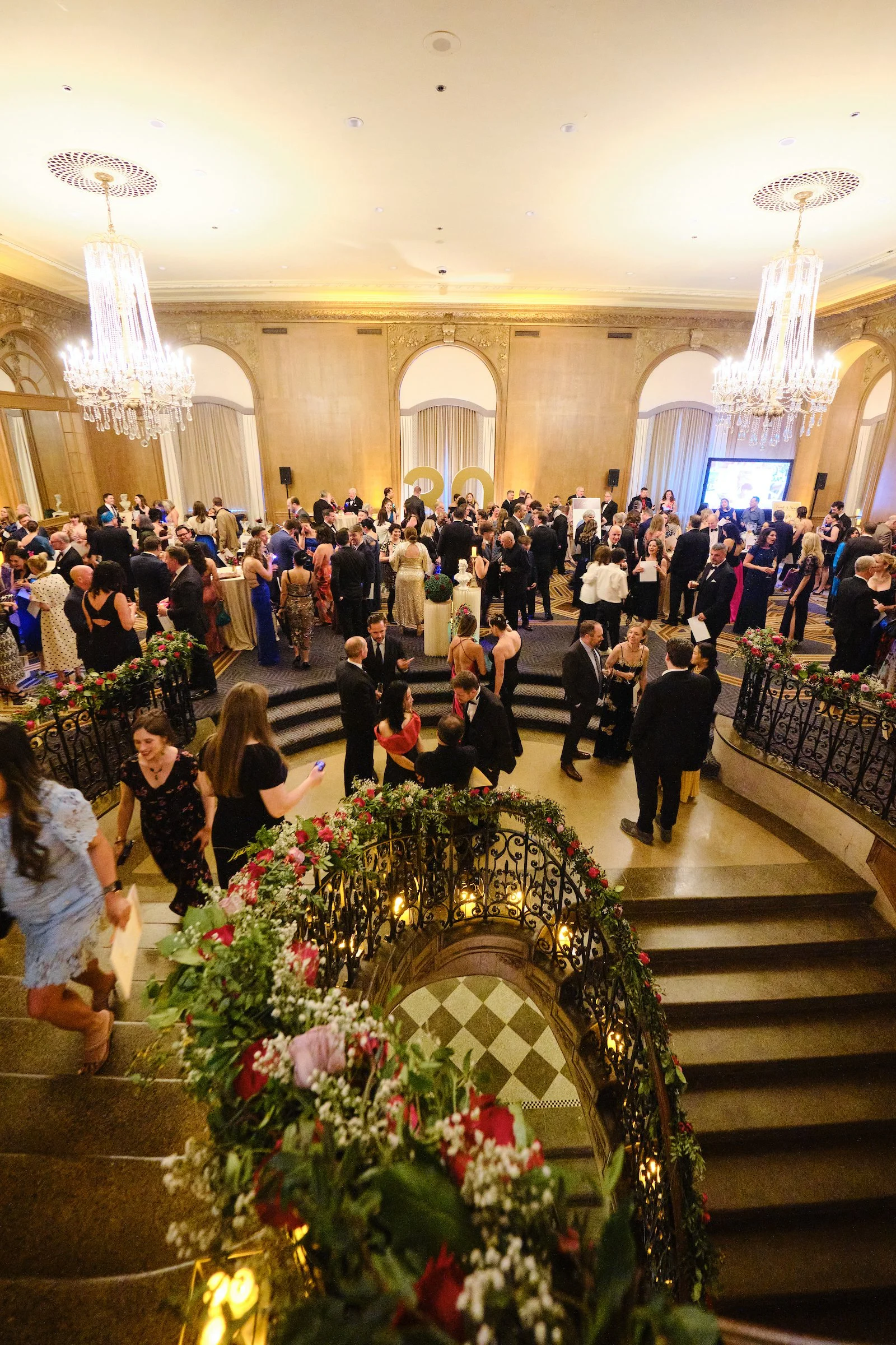 A large ballroom filled with people in formal attire attending an event, with elegant chandeliers, floral decorations, and a staircase in the foreground.