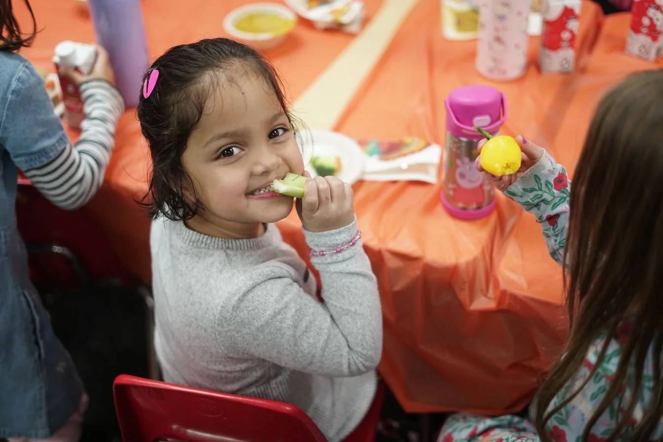 A young girl with dark hair and a pink hair clip smiling while eating a cucumber slice.
