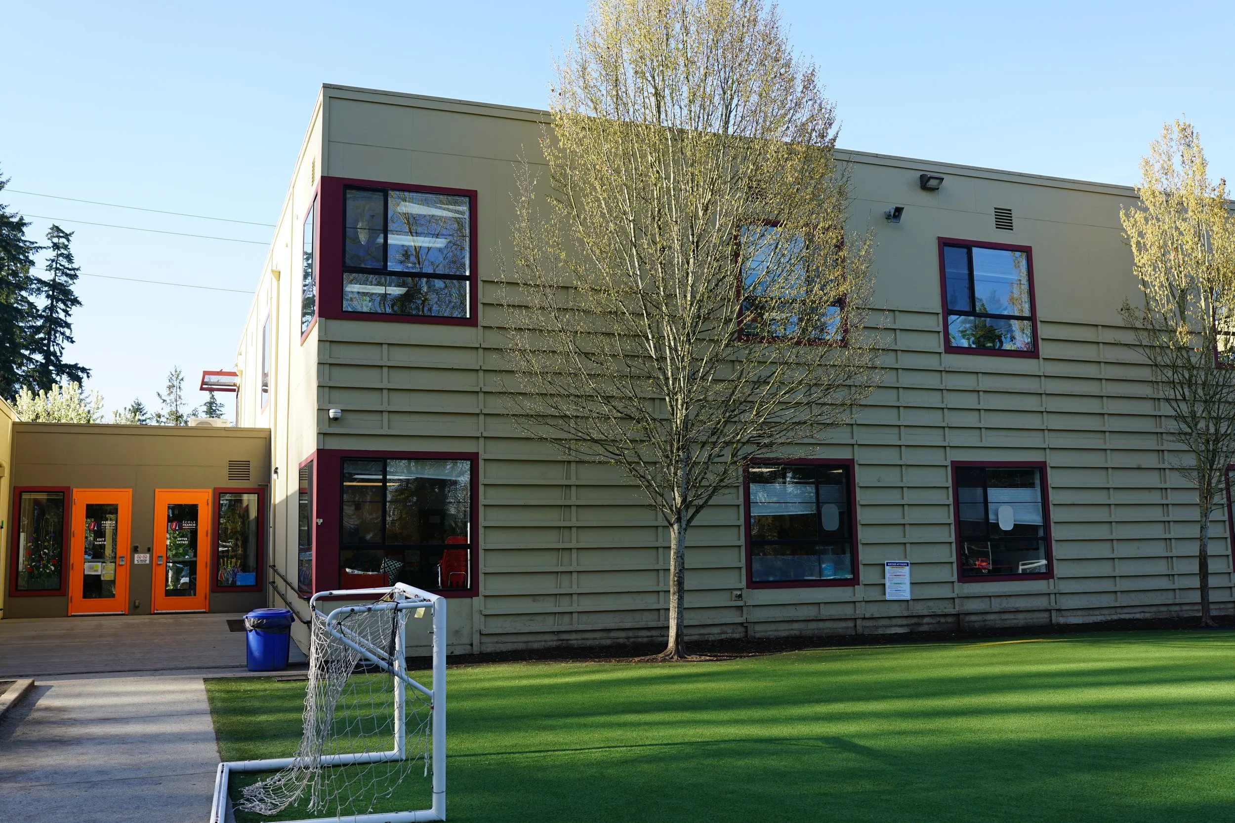 A school building with tan siding and red-trimmed windows, orange doors, surrounded by green grass, trees, and a small soccer goal in the foreground, under a clear blue sky.