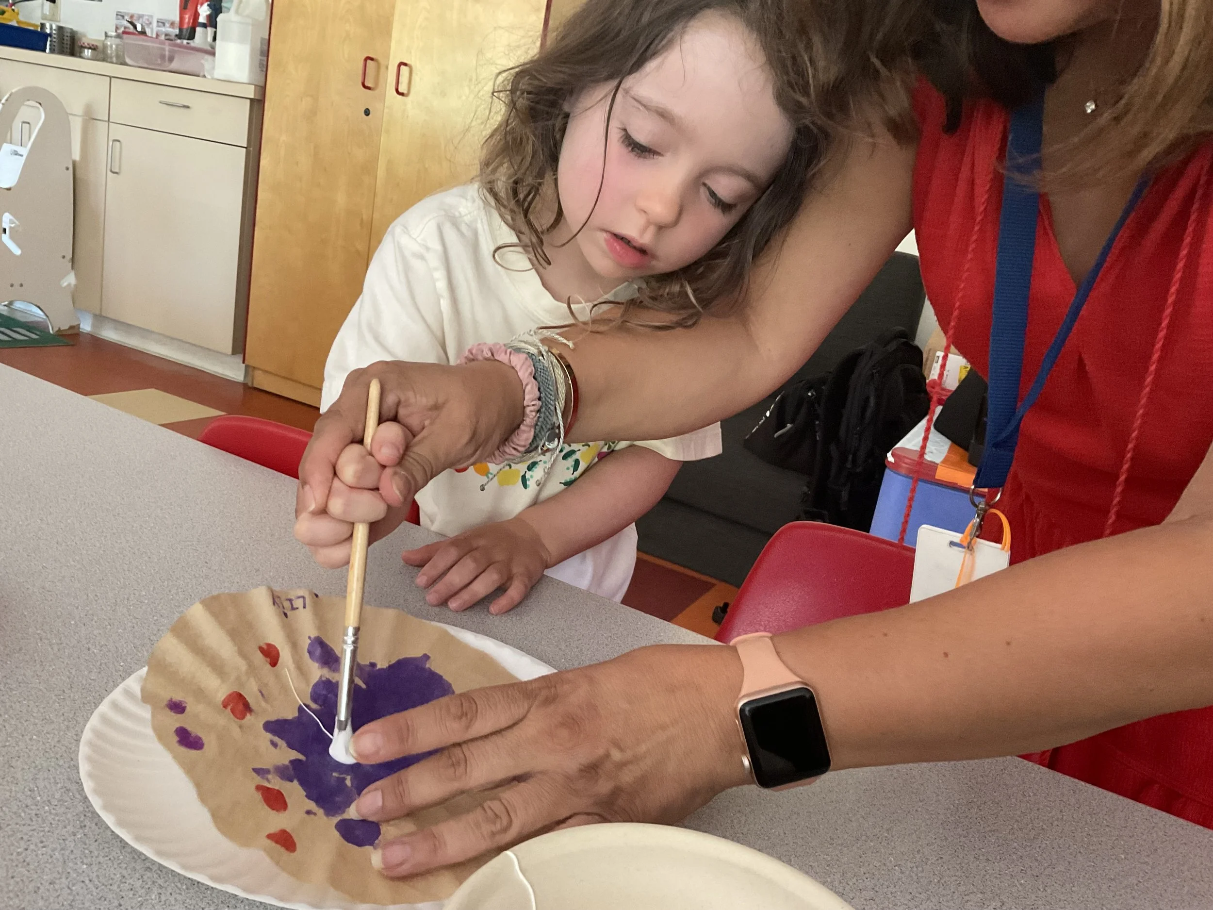 A young girl with curly hair painting on a paper plate with purple and red paint, assisted by an adult wearing a red shirt.