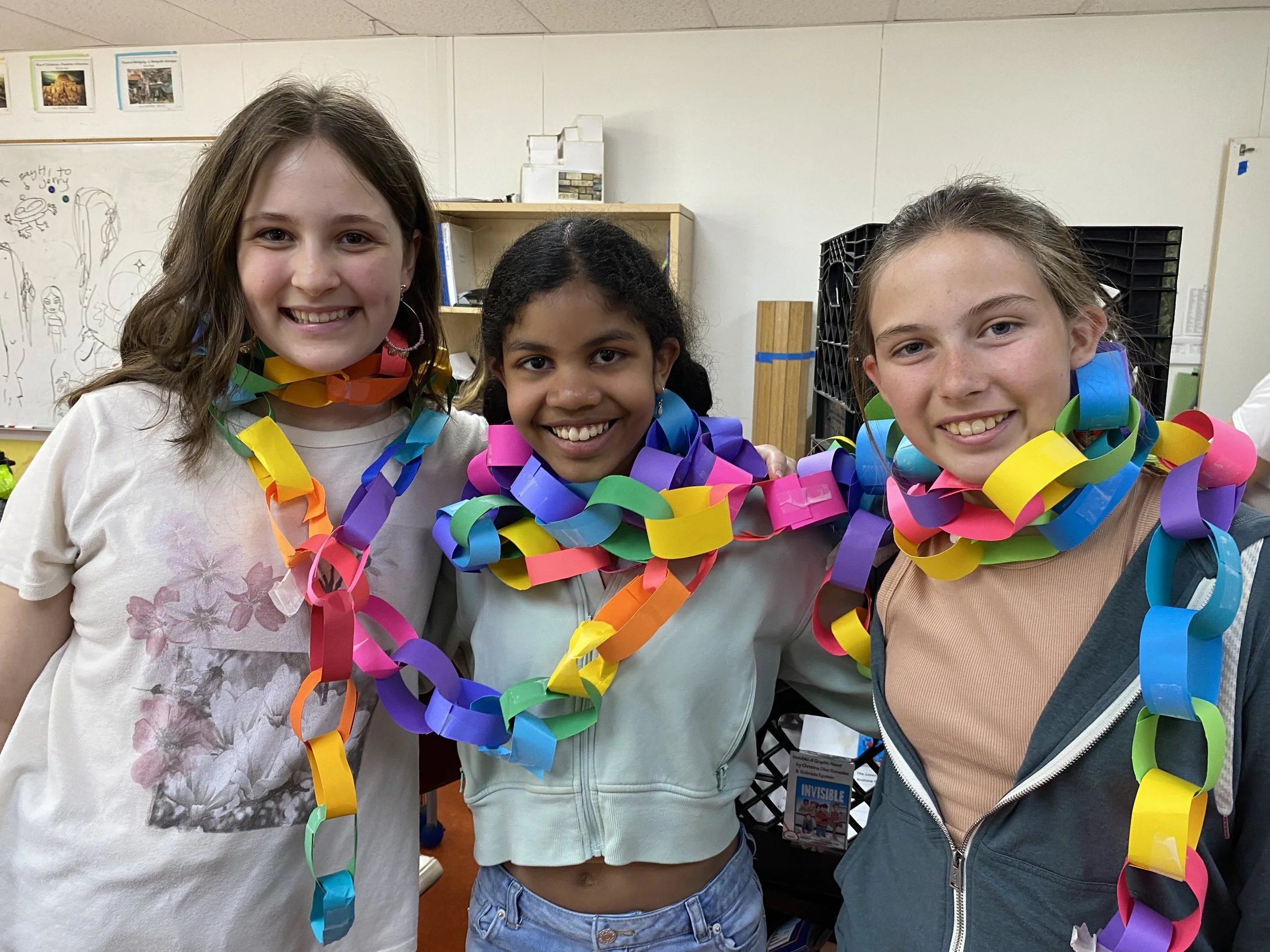 Three young girls smiling and wearing colorful paper chains around their necks in a classroom.