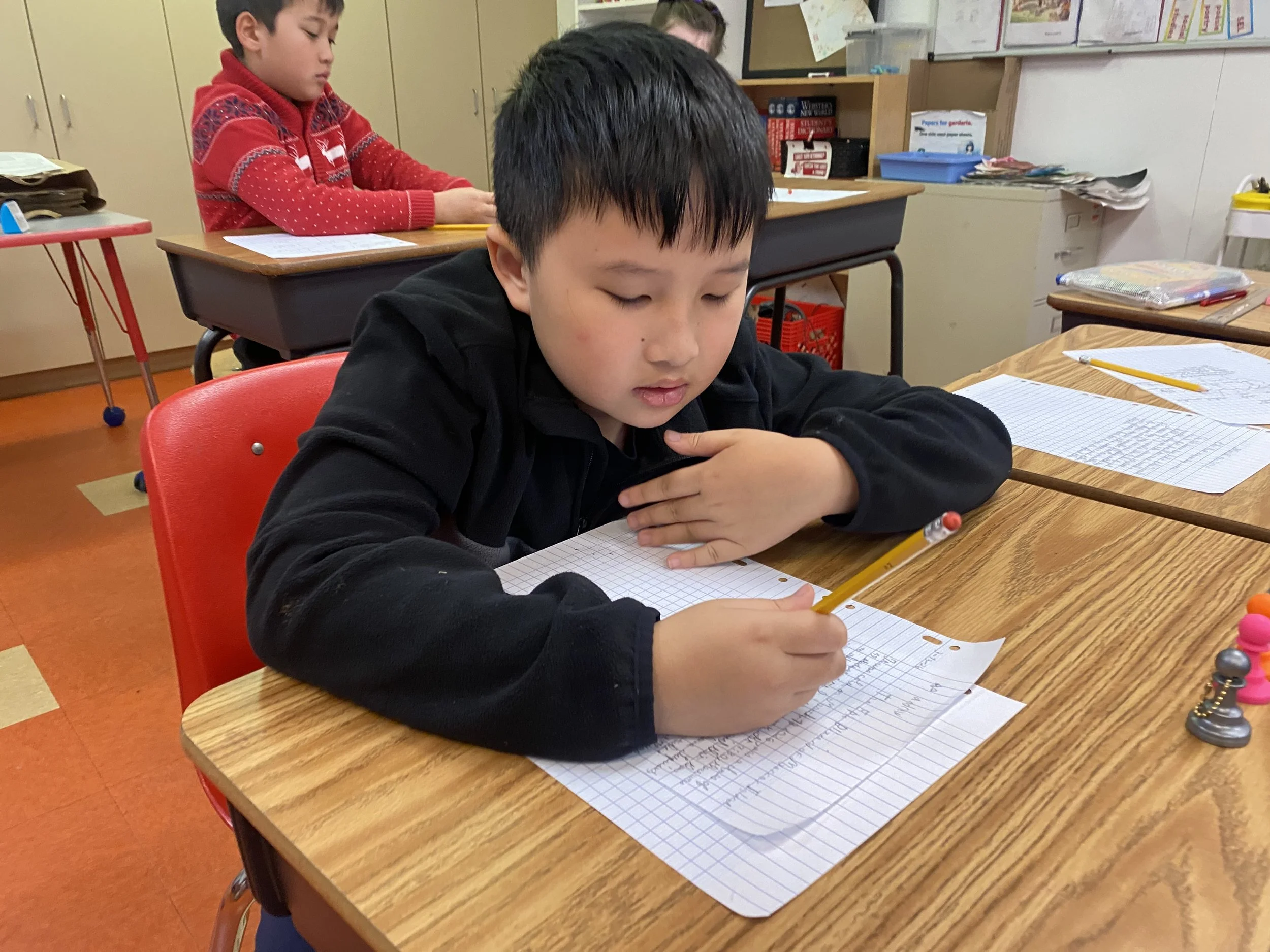 A young boy sitting at a school desk writing in a notebook with a yellow pencil, with a girl in a red sweater sitting behind him in a classroom.