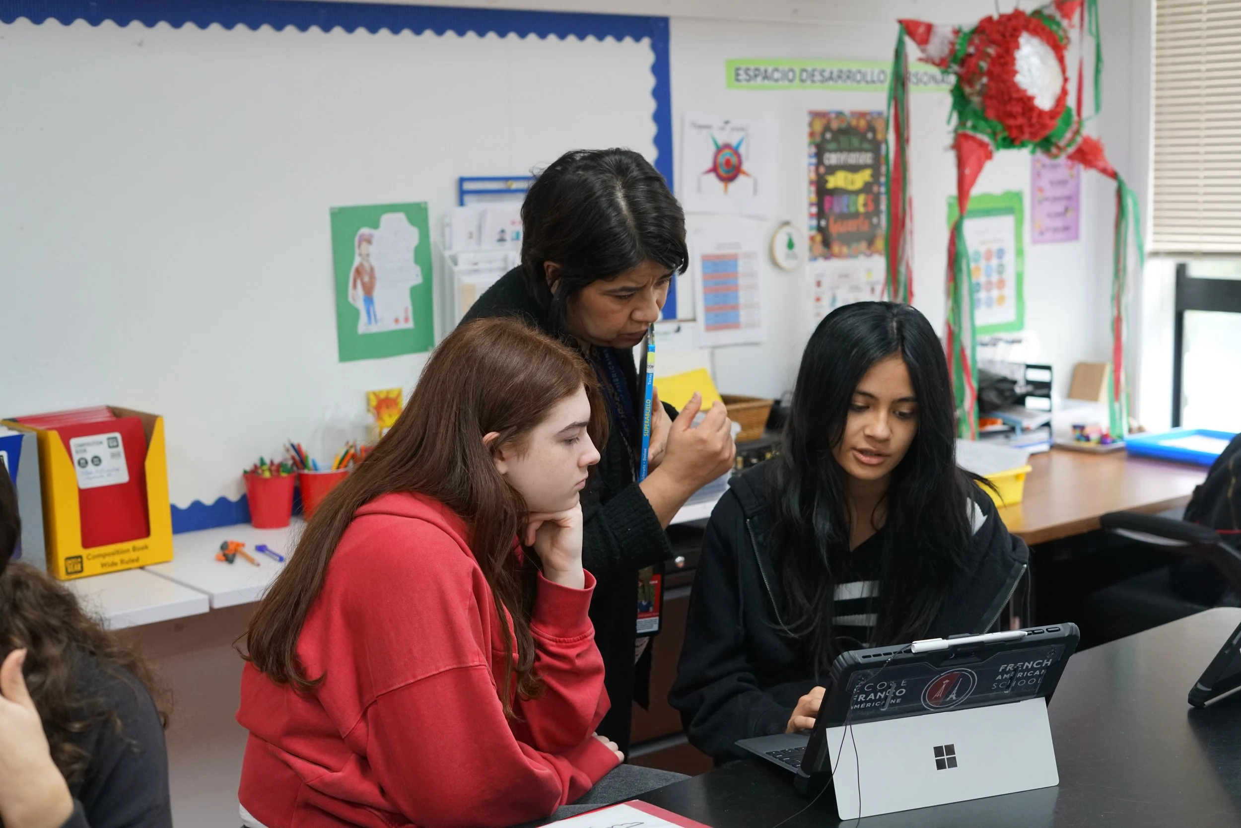 A teacher and three students working together at a classroom table, with educational posters and decorations on the wall behind them.