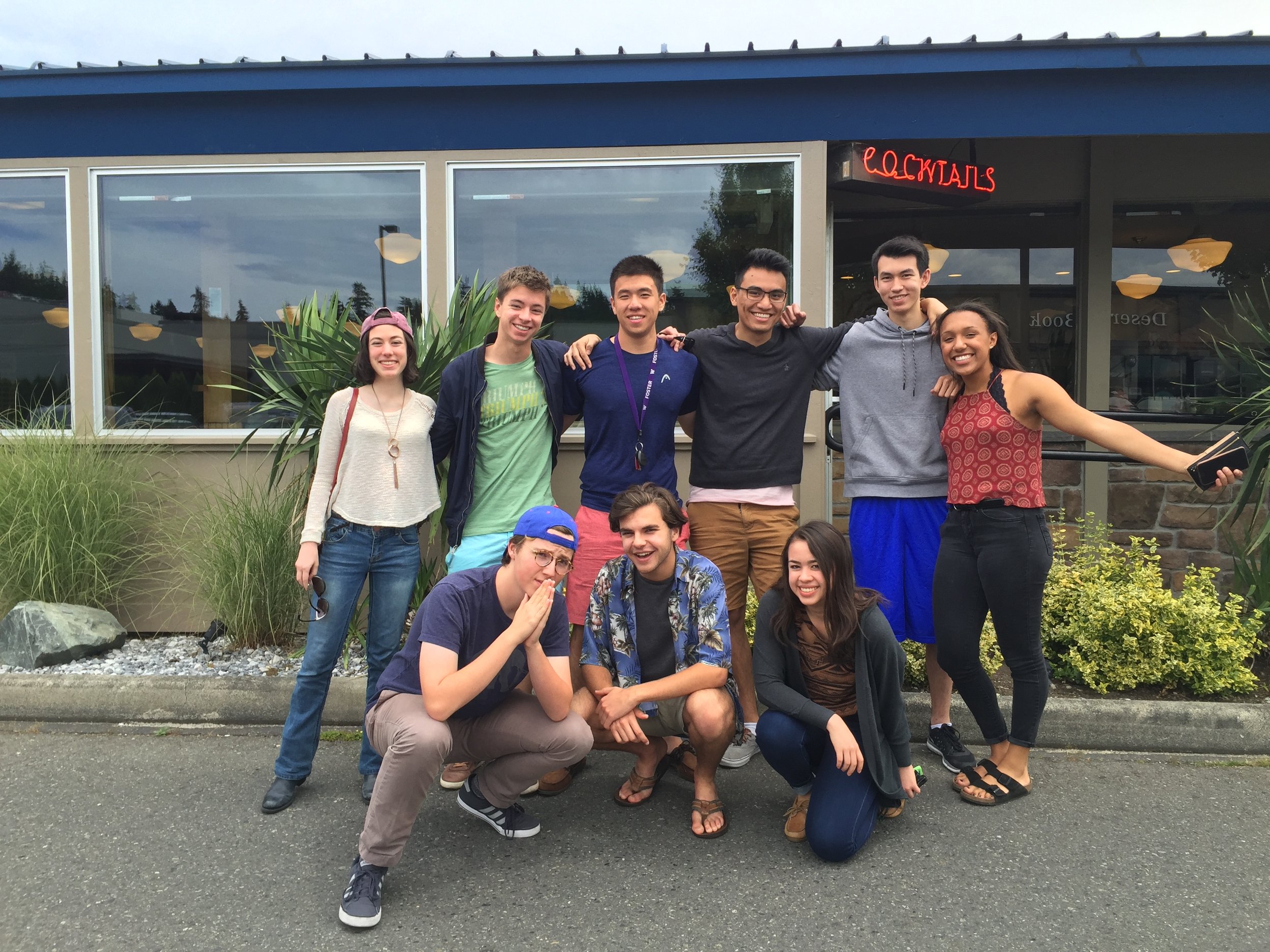 Group of seven young adults smiling and posing for a photo outside a building with a neon sign that reads 'cocktails'; they are standing and squatting on a sidewalk with some greenery and rocks in the foreground.