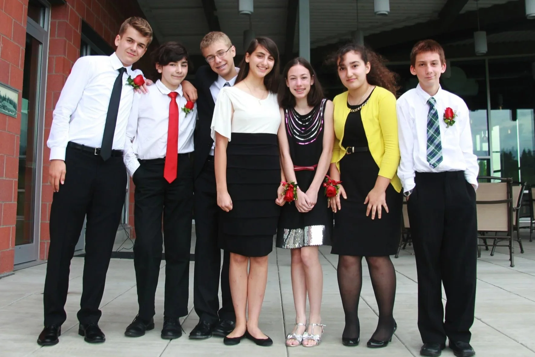 Group of eight teenagers dressed in formal attire, standing outside a building, smiling, with some wearing corsages and boutonnières.