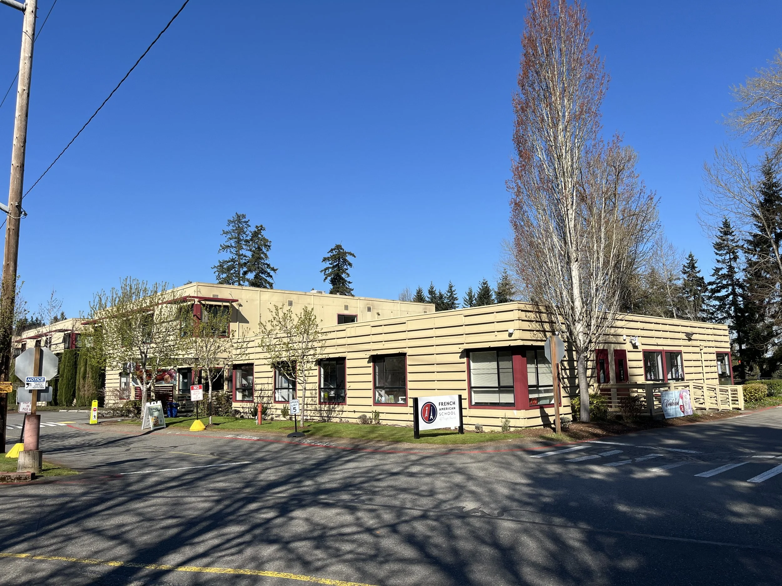 A single-story building with large glass windows and a beige wooden exterior. There is a sign reading French American School at the front. Several small trees and bushes are outside, along with parking spaces and a street in front. Tall trees are visible in the background under a clear blue sky.
