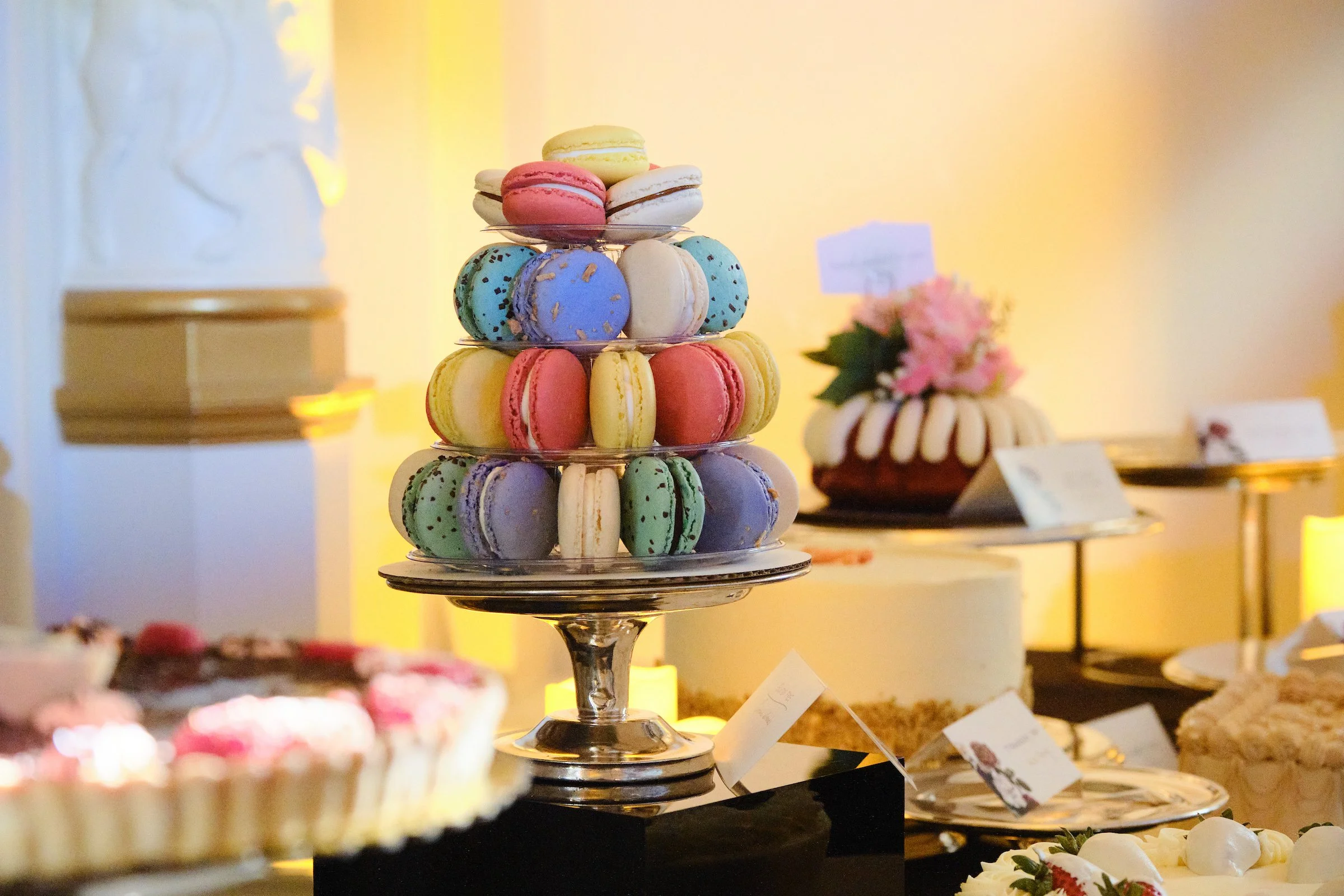 A multi-tiered metal dessert stand filled with colorful macarons including yellow, pink, blue with black specks, white, and beige, at a dessert display or buffet.