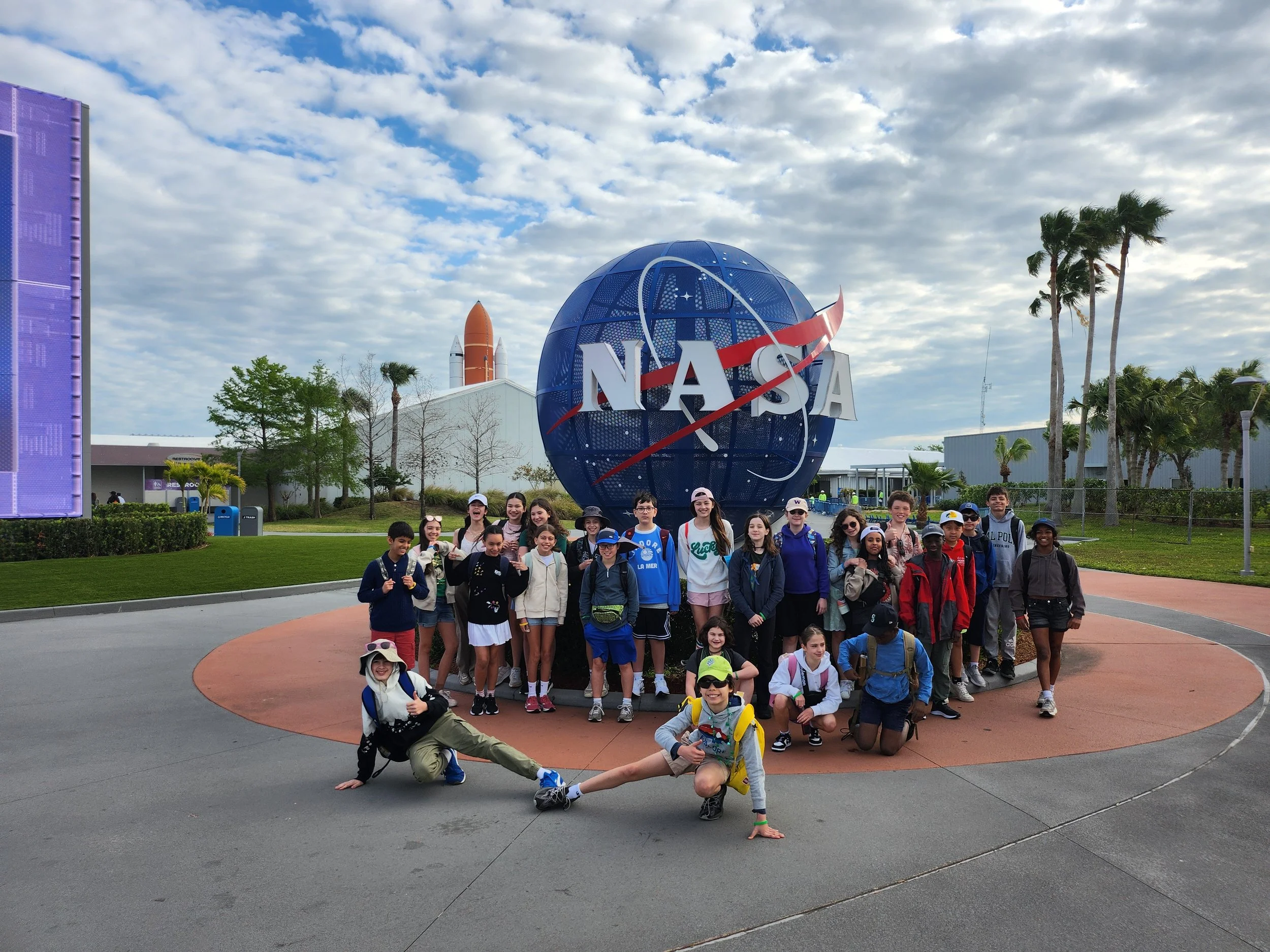 A group of children and adults posing in front of a large NASA logo at a space center, with a blue sky and scattered clouds overhead, and palm trees in the background.