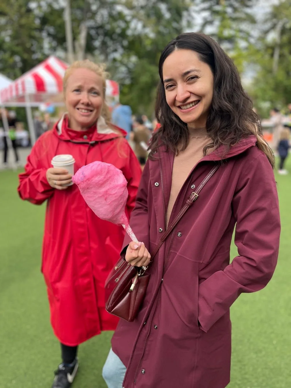 Two women smiling at an outdoor event; one holding pink cotton candy, the other holding a coffee cup, with tents and other people in the background.