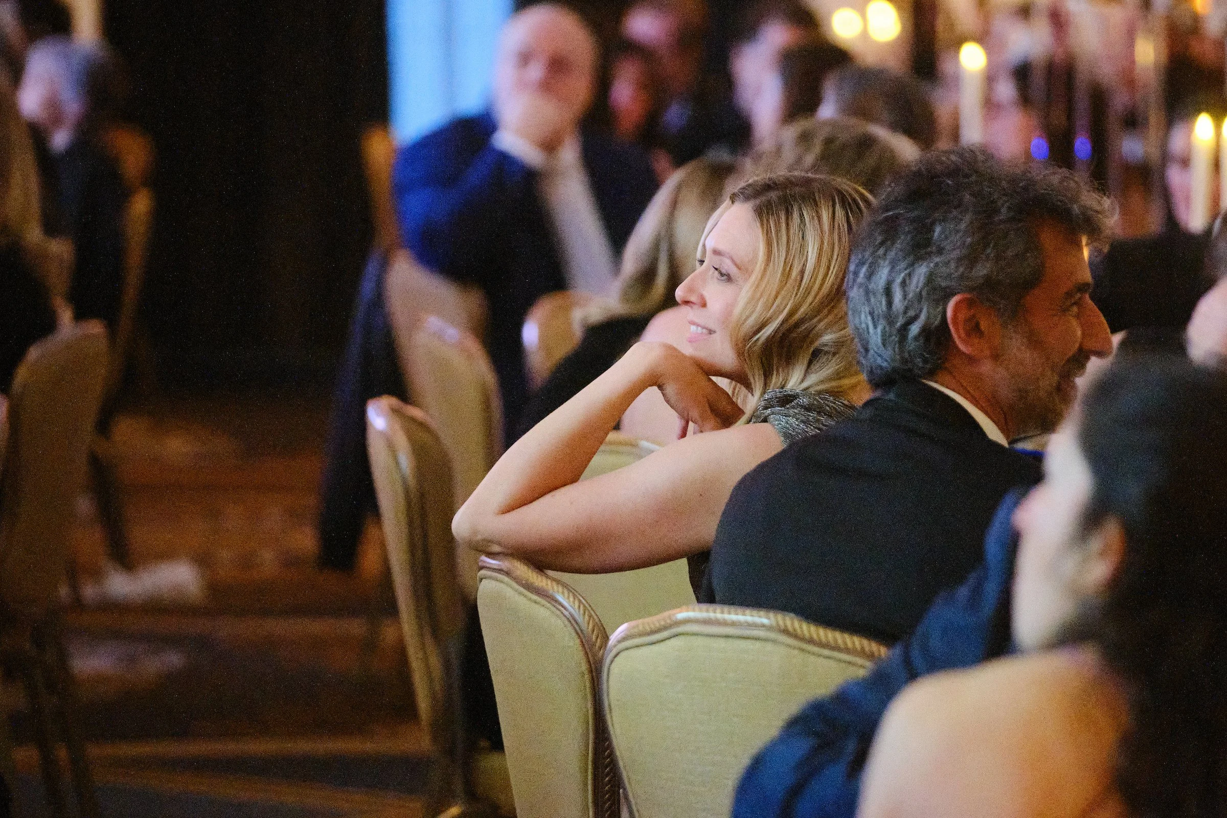 People attending a formal event or banquet, seated at tables, engaging in conversation, with warm ambient lighting.