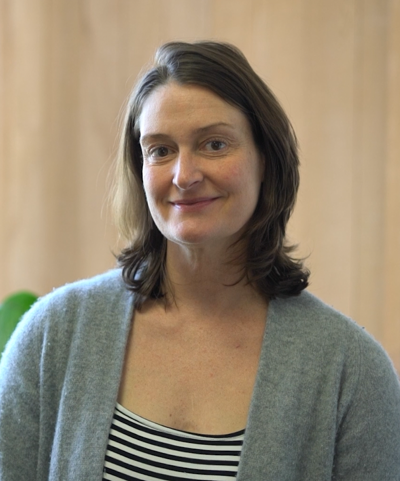 A woman with shoulder-length brown hair and fair skin, smiling, wearing a grey cardigan over a black and white striped top, standing indoors with a beige background.