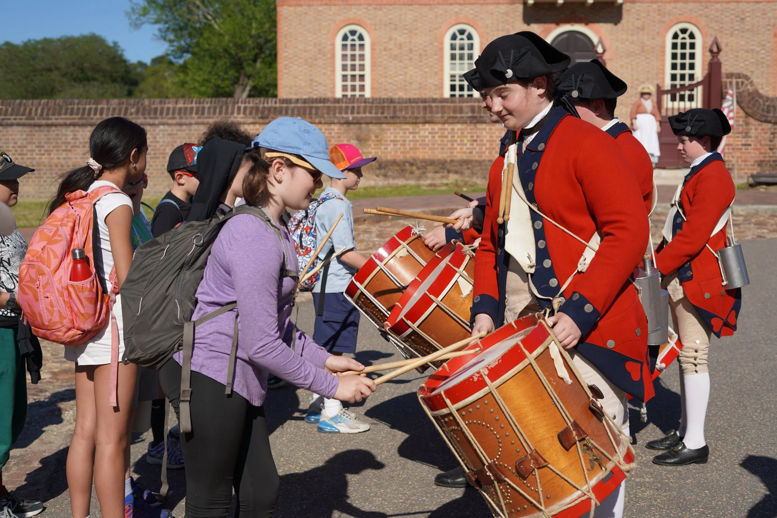 A group of children standing in line, being addressed by individuals dressed in Revolutionary War military uniforms, who are holding drums. The children are wearing backpacks and hats, and appear to be visiting a historical site with a brick building in the background.
