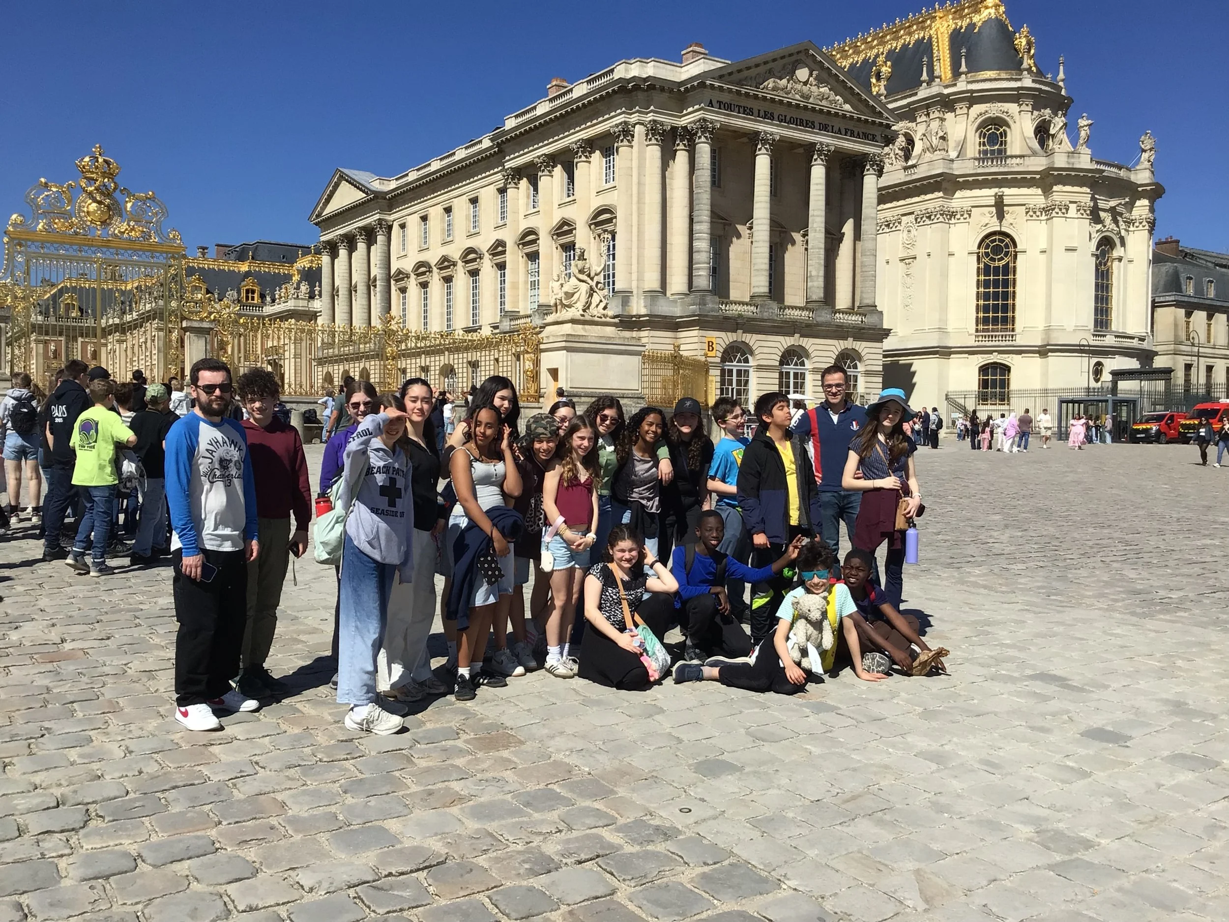 A group of tourists, including children and adults, standing in front of a grand, historic building with classical architecture and gold accents, possibly the Palace of Versailles in France.