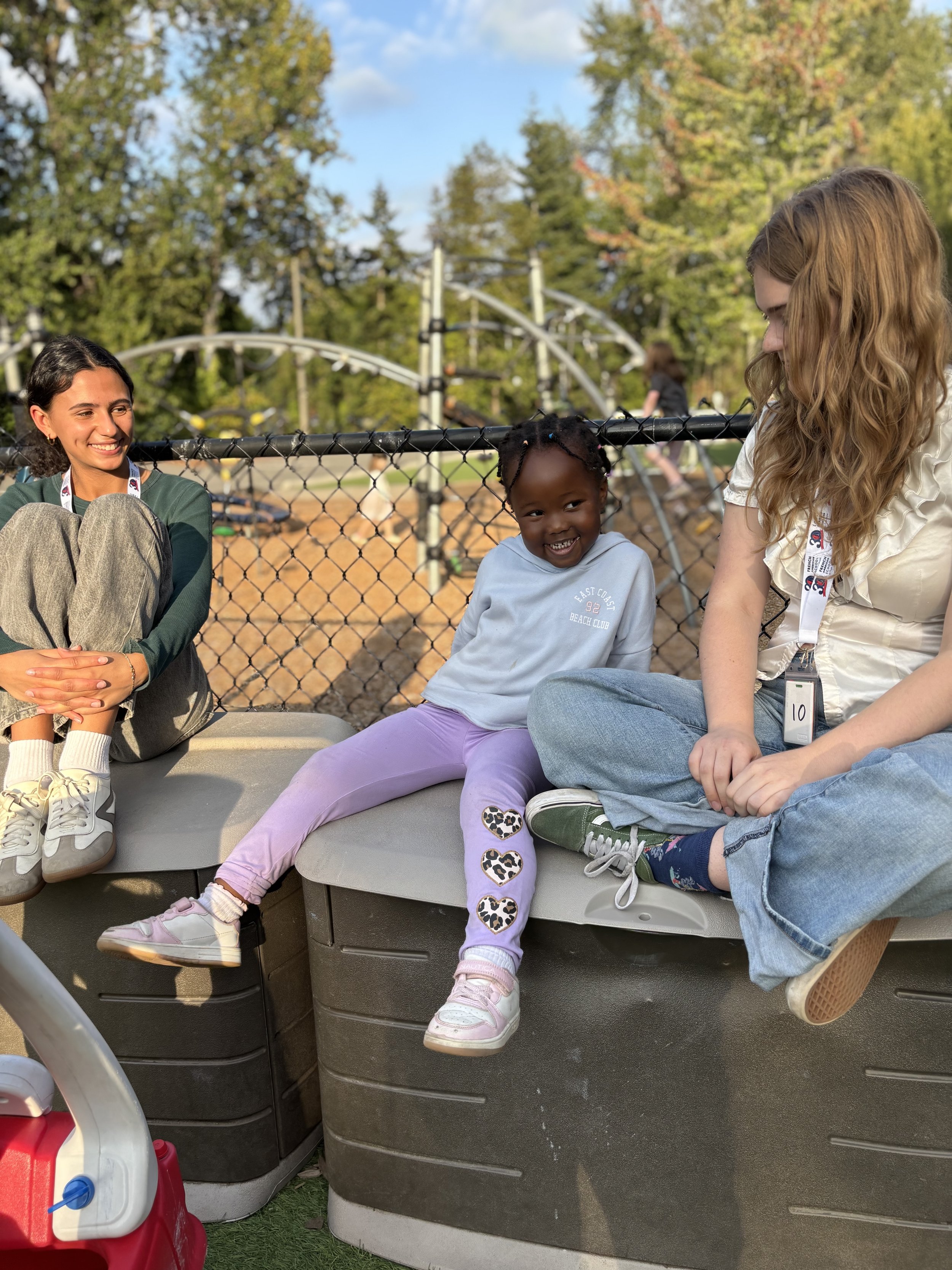 Three women and a girl sitting outdoors near a fence, smiling and talking, with a roller coaster and trees in the background.