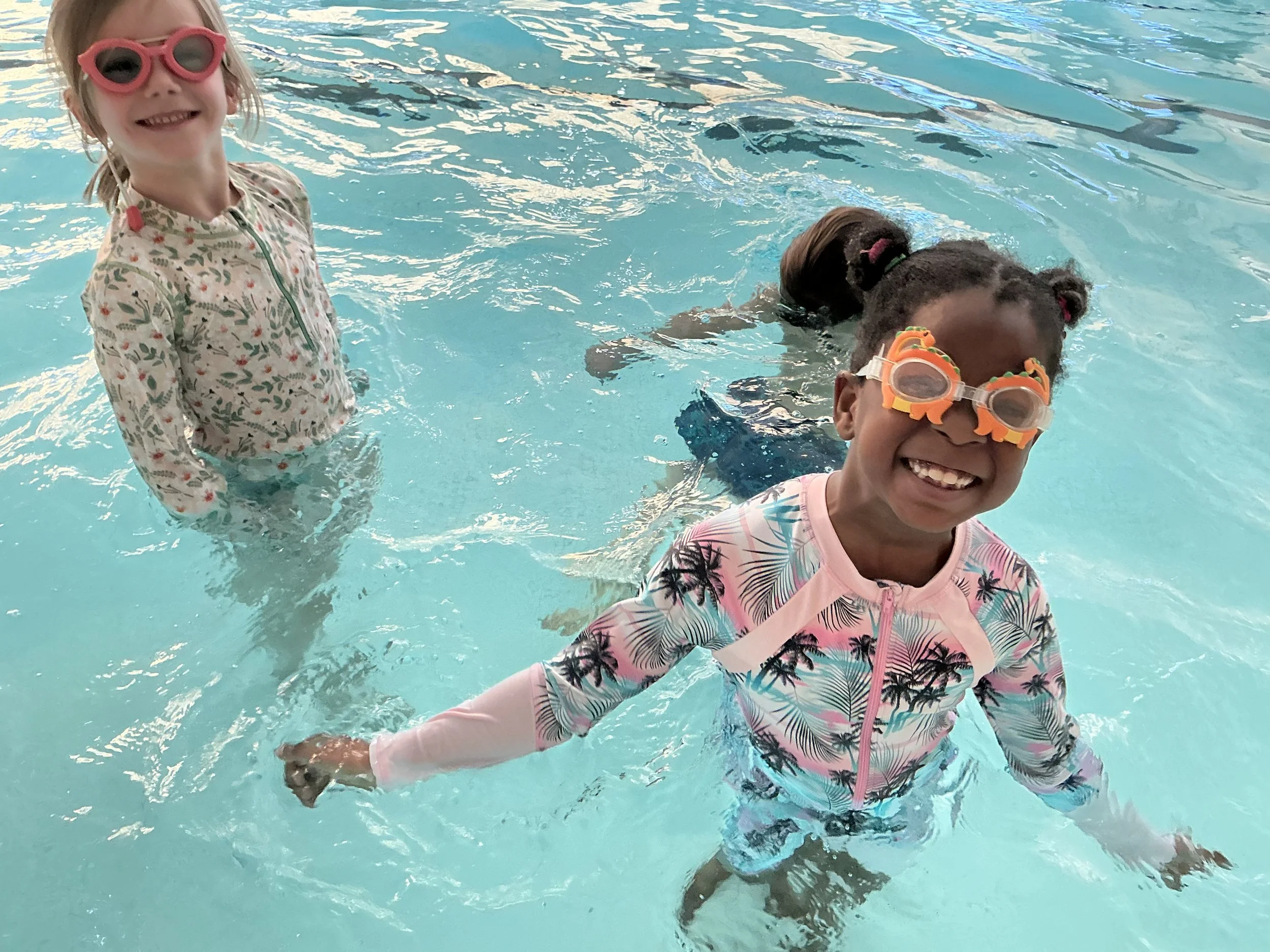 Two children smiling and playing in a swimming pool, wearing colorful swimwear and goggles.