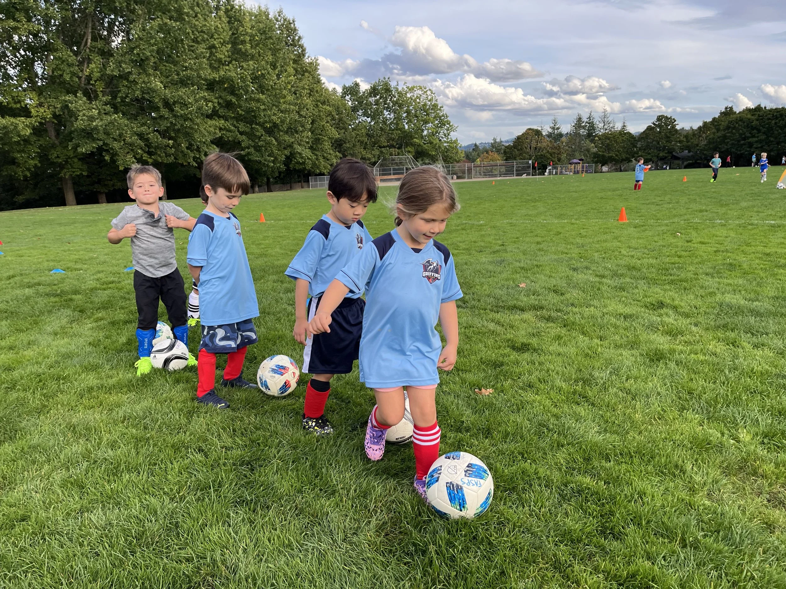 Young children standing in a line on a grassy field, each with a soccer ball at their feet, wearing sports uniforms, during a soccer practice on a cloudy day.