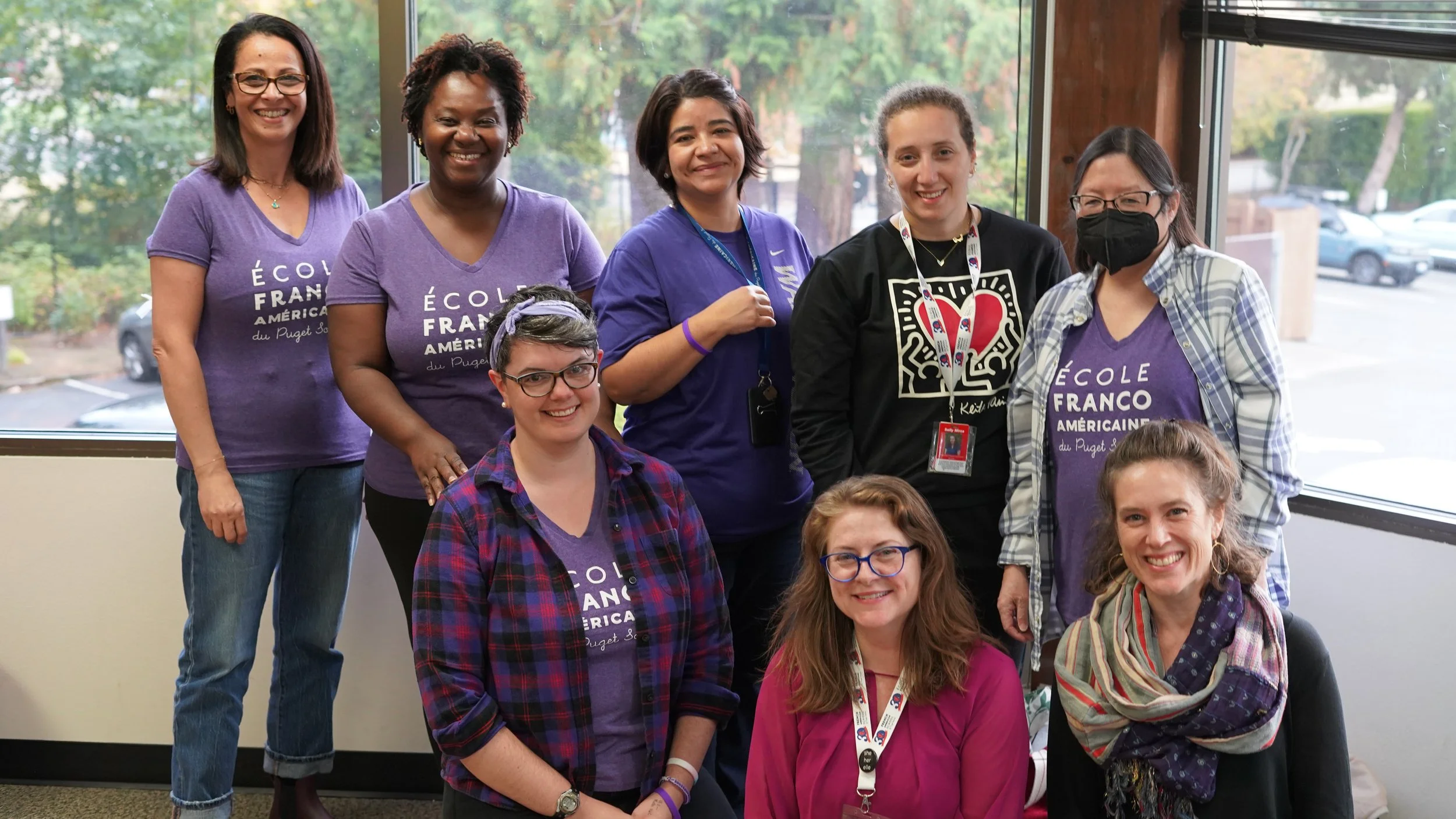 Group of diverse women and girls smiling indoors, some wearing purple t-shirts with text, standing in front of a window with trees visible outside.
