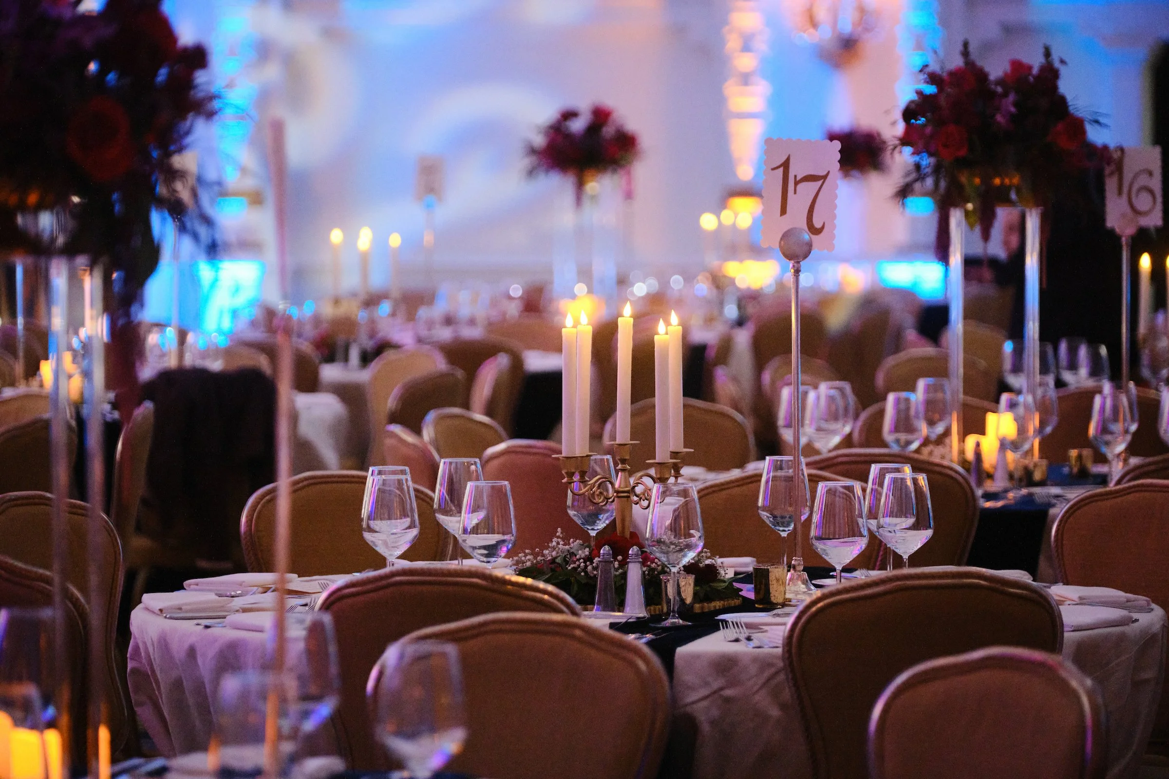 A banquet hall decorated with floral centerpieces and candles on tables, with a backdrop of colorful lighting, set for an event or celebration.